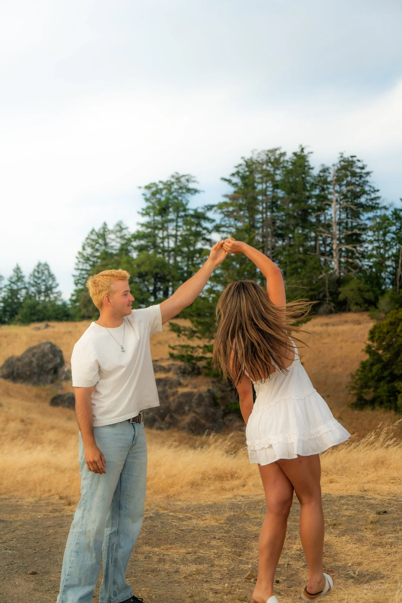 A young man and woman dancing outdoors in a grassy, wooded area with trees in the background, holding hands and spinning.