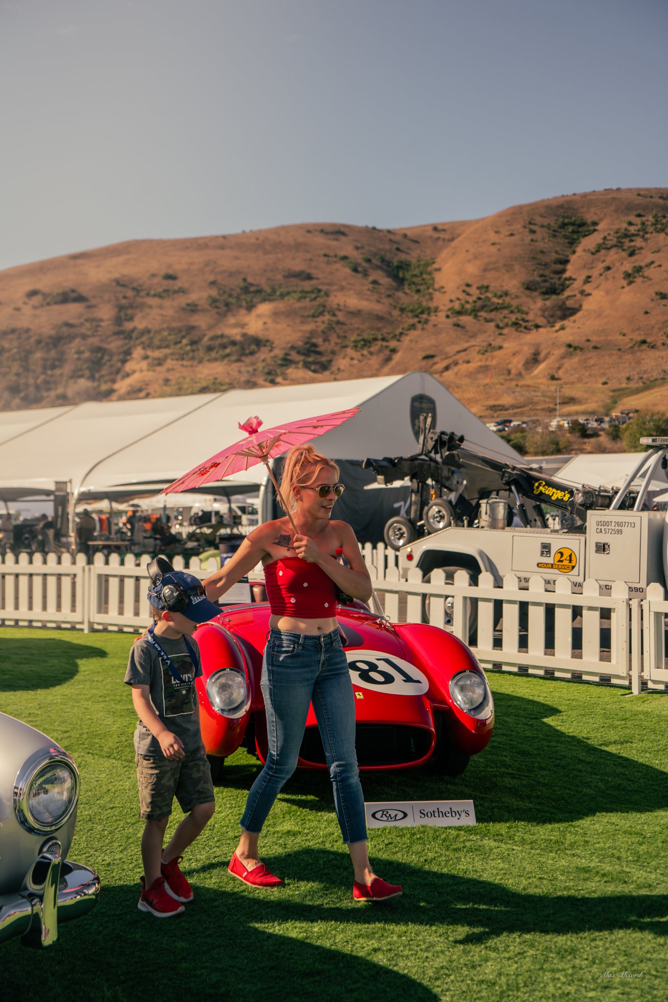 A woman holding a pink umbrella walking past a red vintage race car at an outdoor car auction, with a young boy walking beside her. There are cars and a tent in the background and mountains in the distance.
