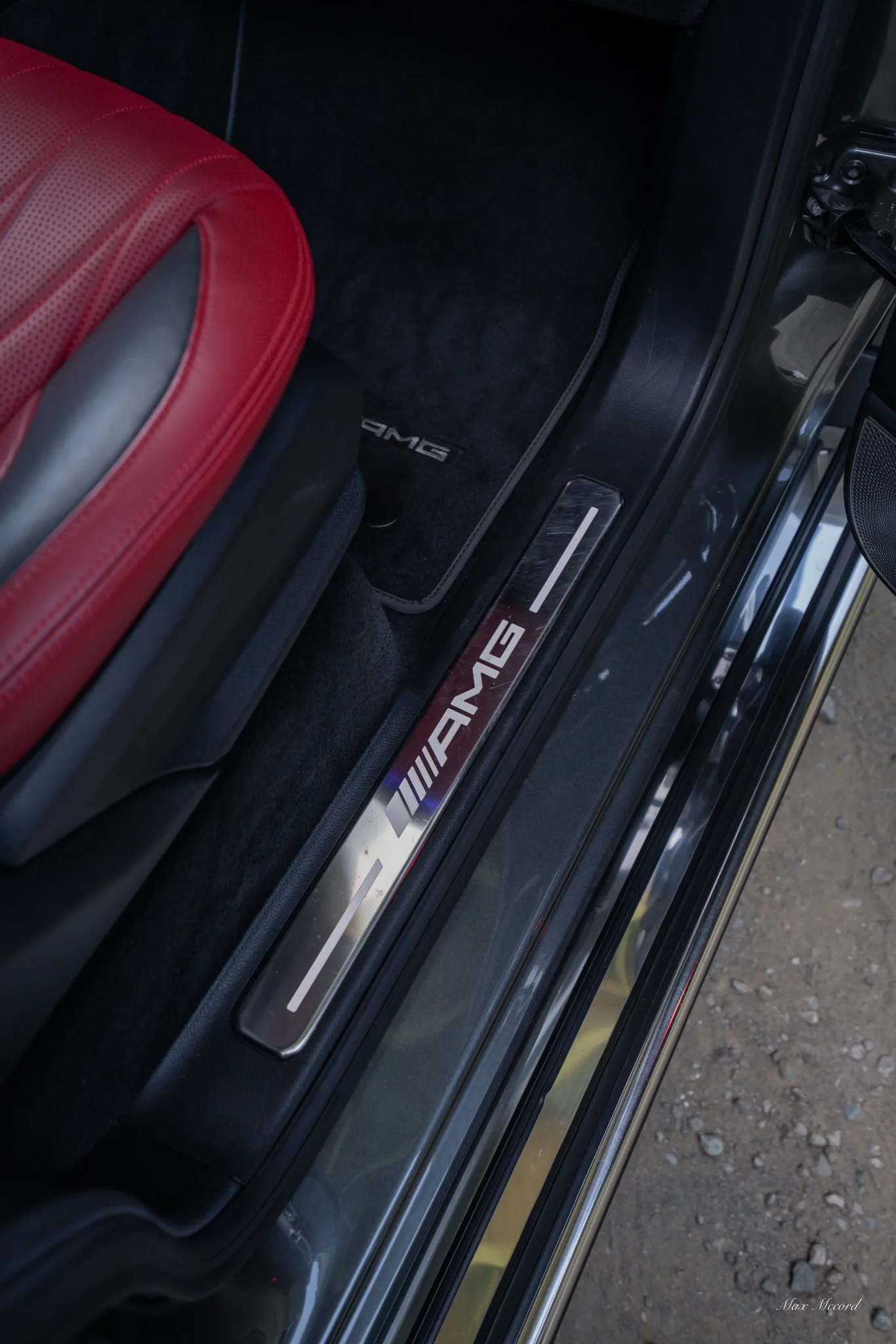 Close-up of car door sill with AMG logo, red leather seat, black floor mat with AMG branding, and black car interior.