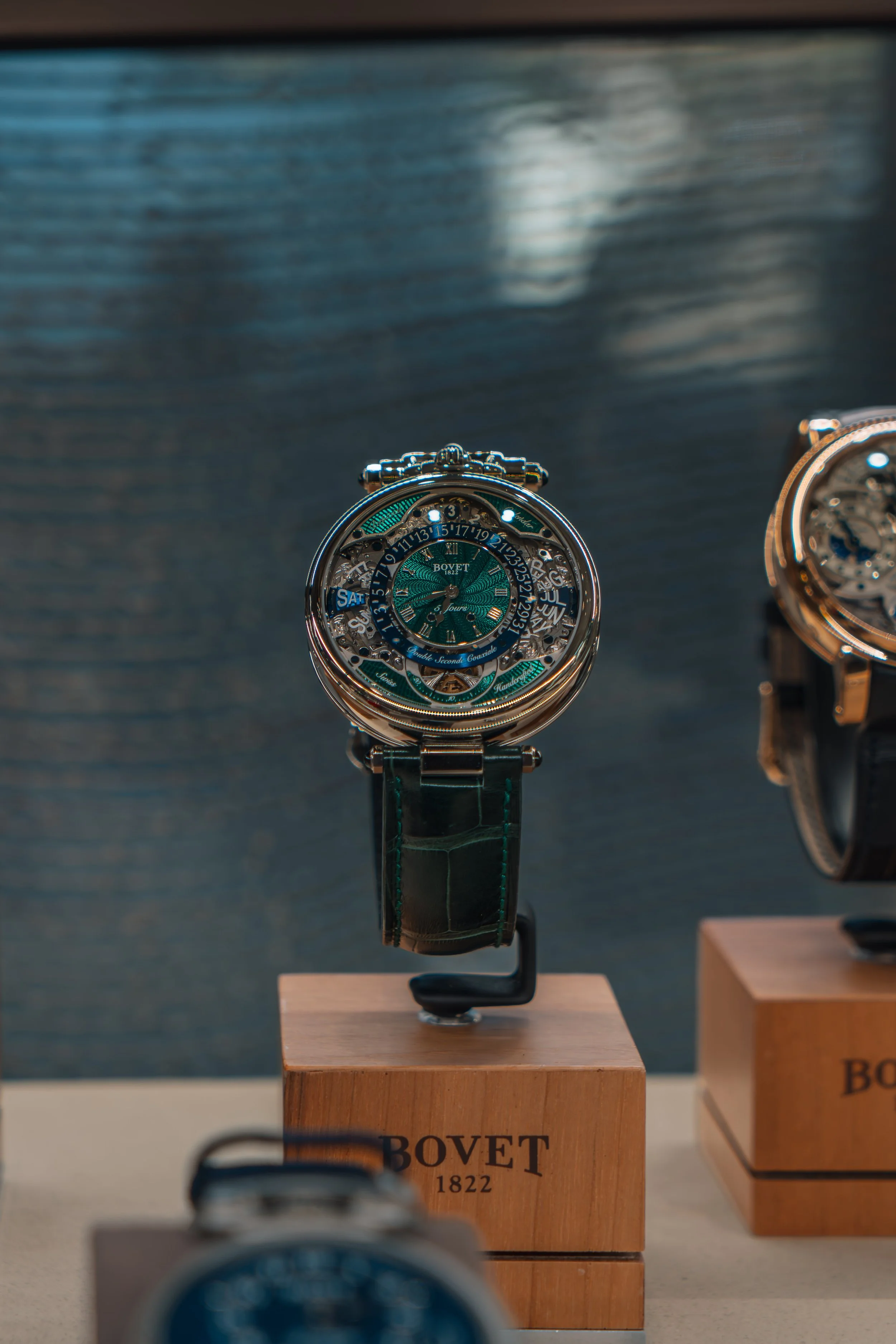 A luxury wristwatch displayed on a wooden stand with the brand Bovet 1822, featuring a complex dial with multiple functions and a green leather strap.