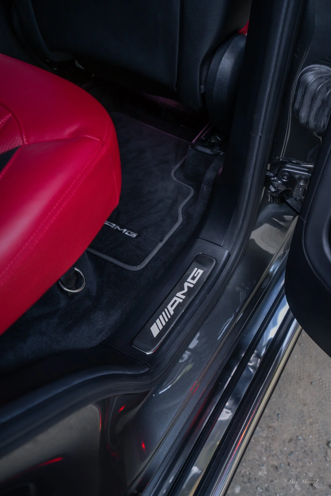 Close-up of the inside of a car showing AMG branded floor mats, a red leather seat, and a door sill with an AMG logo.