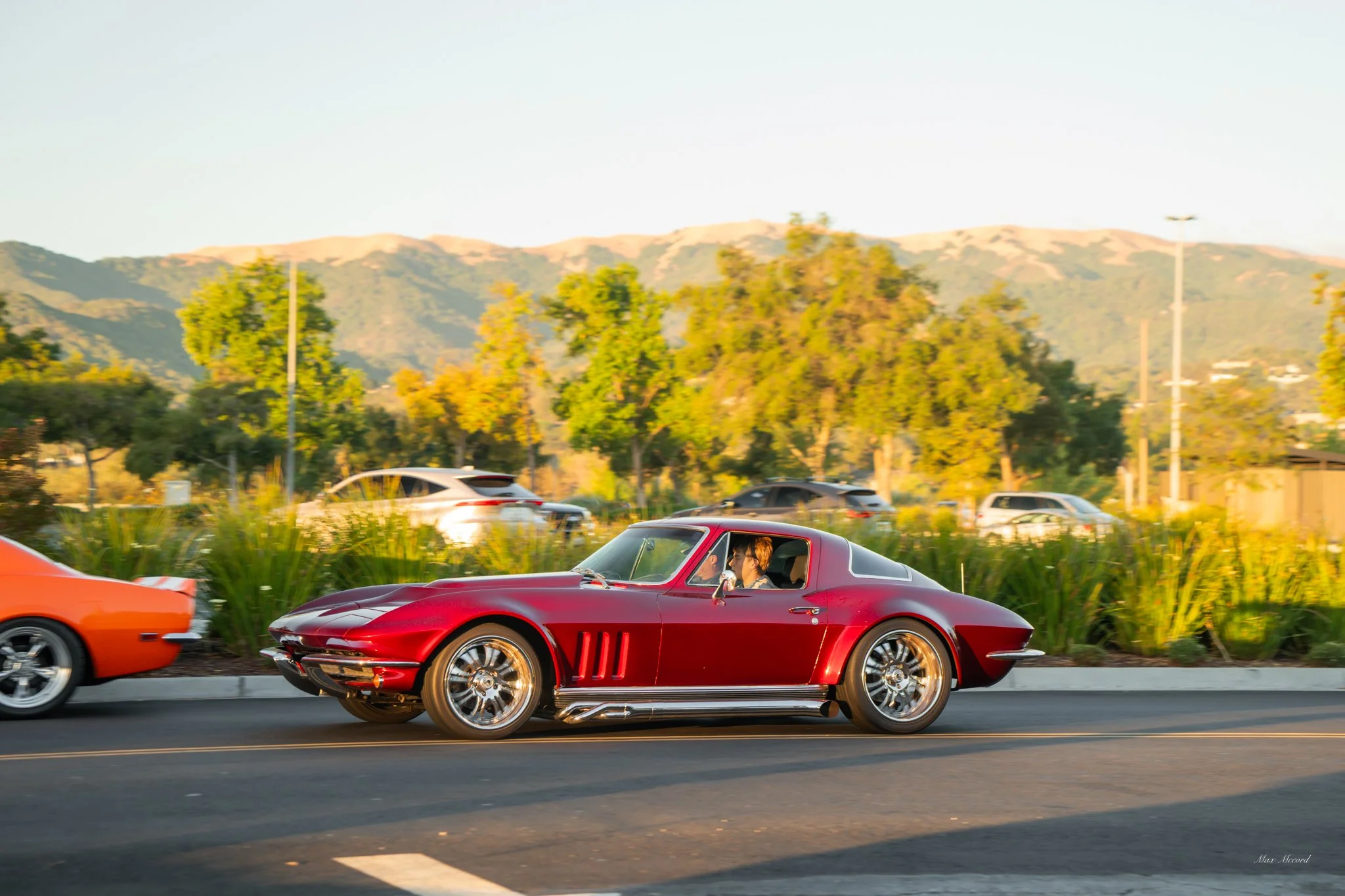 A red vintage sports car being driven on a city street during daytime, with trees and mountains in the background.