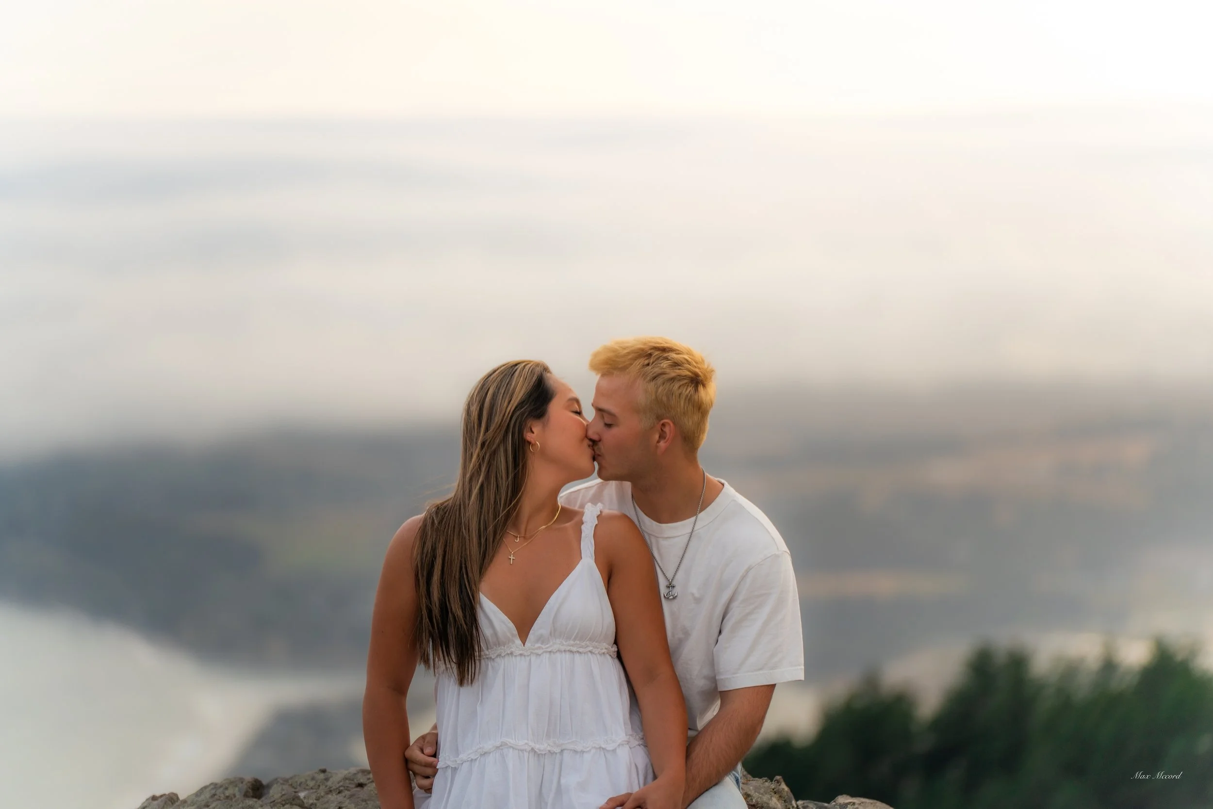 A young couple sharing a kiss outdoors with a scenic landscape in the background.