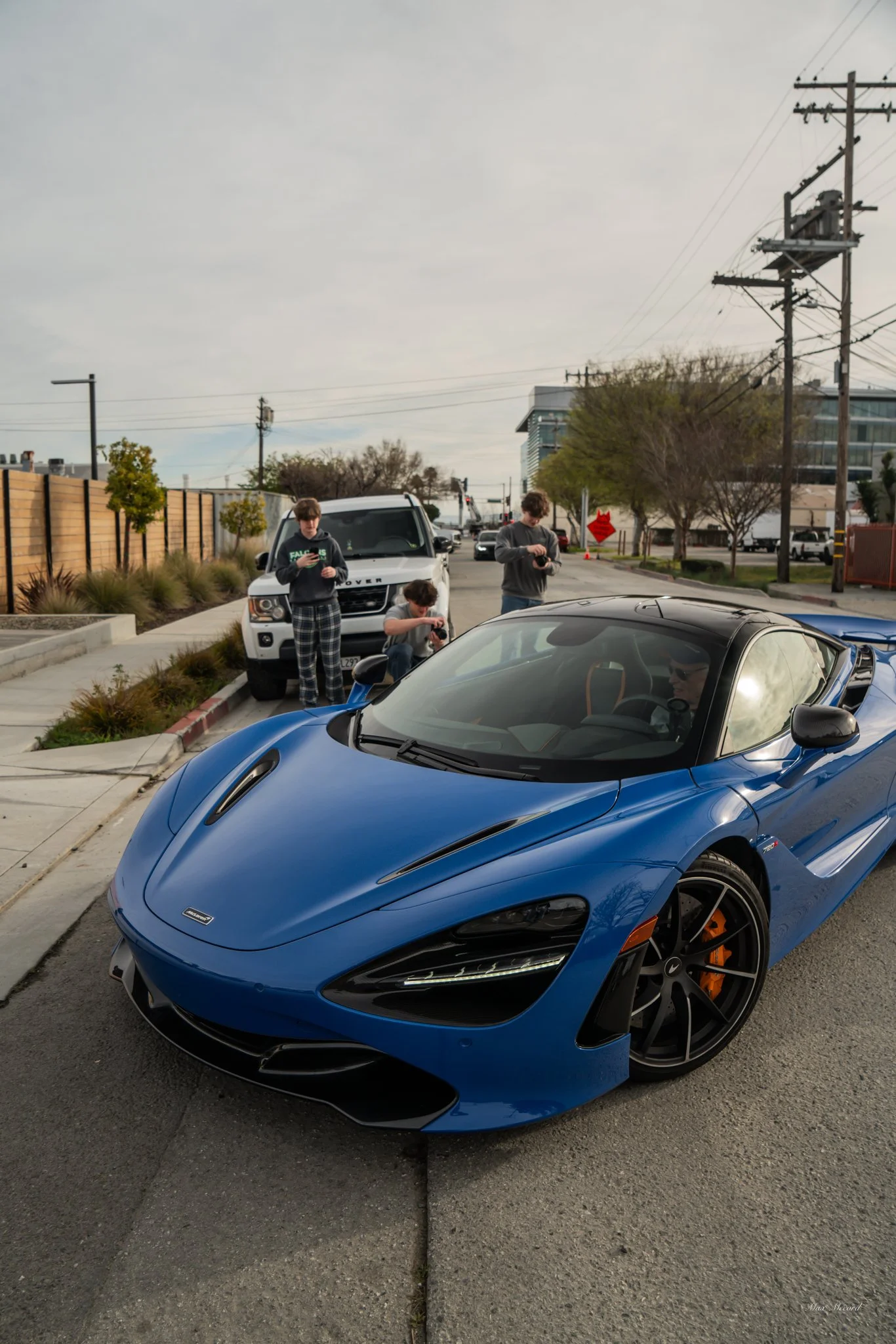 A blue sports car parked on the side of the street with three young men standing and one sitting, all using their phones. A white SUV is parked behind the sports car. The background includes a sidewalk, some trees, utility poles, and buildings.