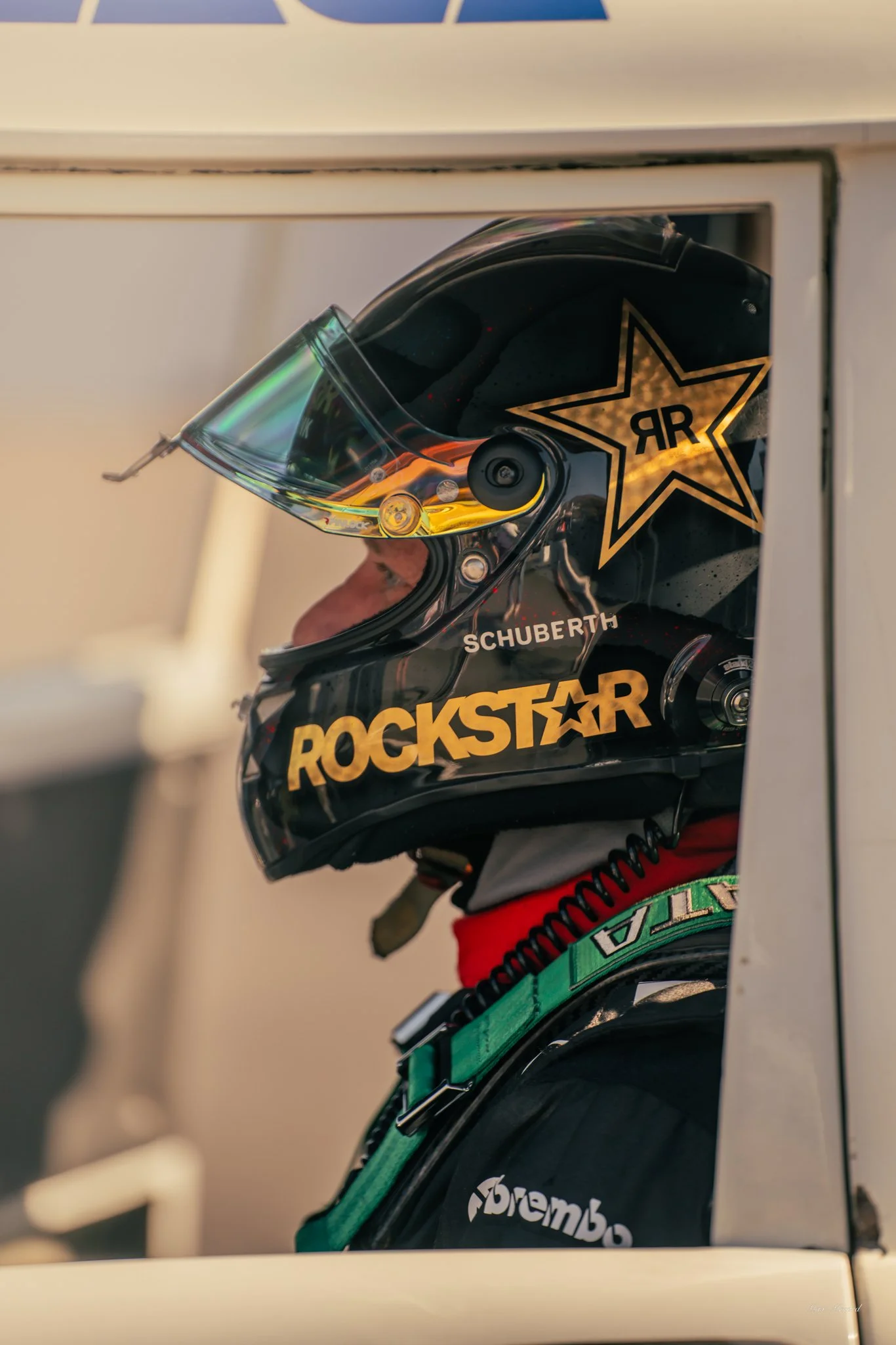 Racing driver in a black helmet with Rockstar and Schuberth logos, seen from the side, inside a vehicle preparing for a race.