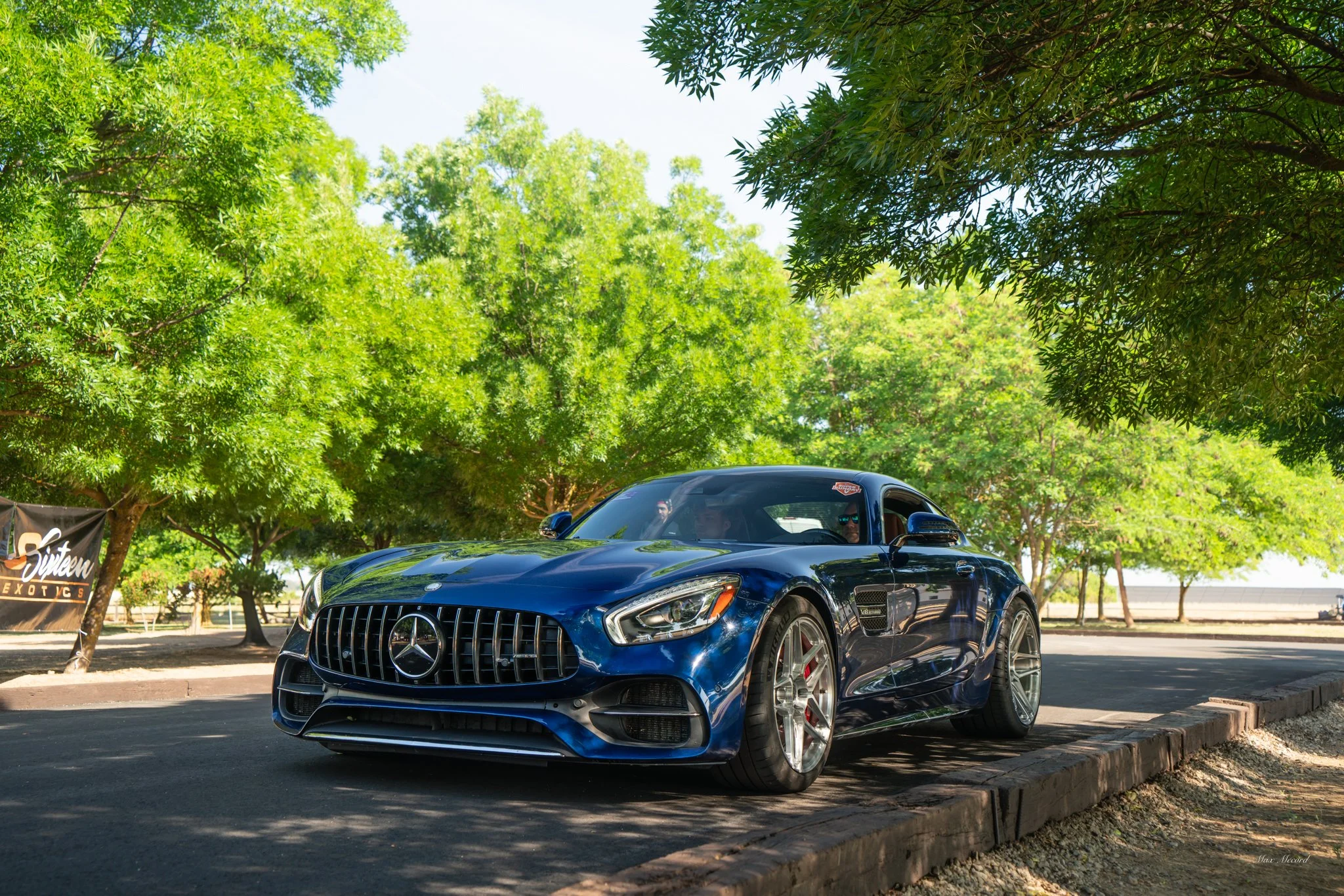 A blue Mercedes-Benz sports car parked on a tree-lined street with lush green trees and a clear sky in the background.