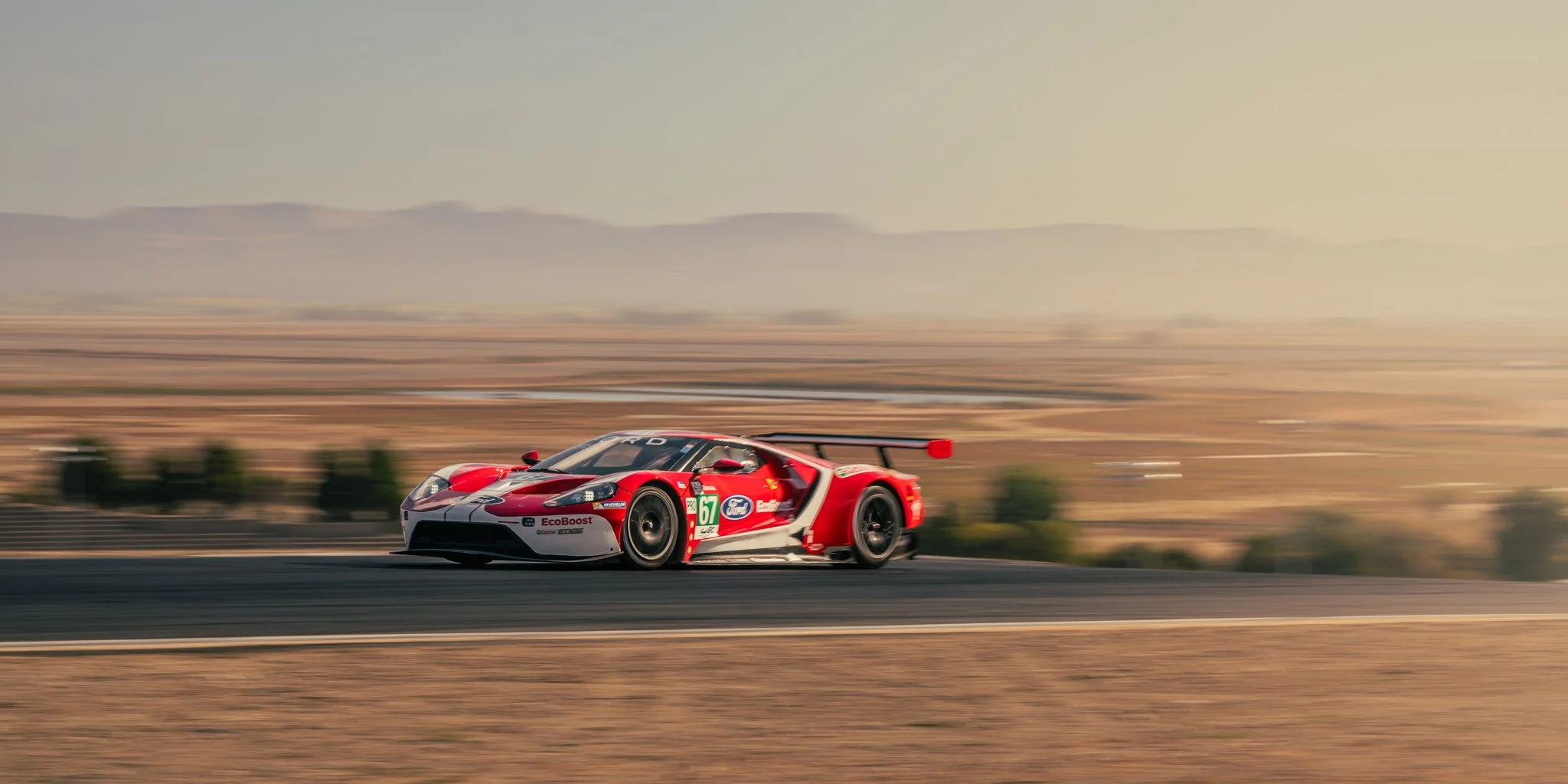 Red and white racing car speeding on a track in a desert landscape with mountains in the background.