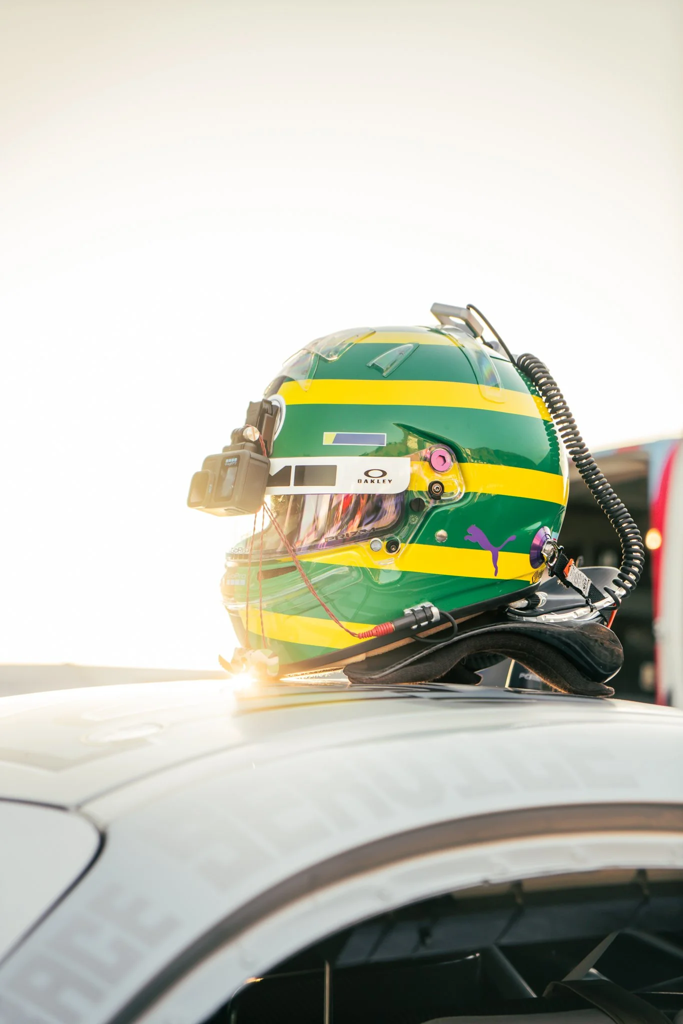 A green and yellow racing helmet with various attachments, placed on top of a white vehicle, with sunlight in the background.