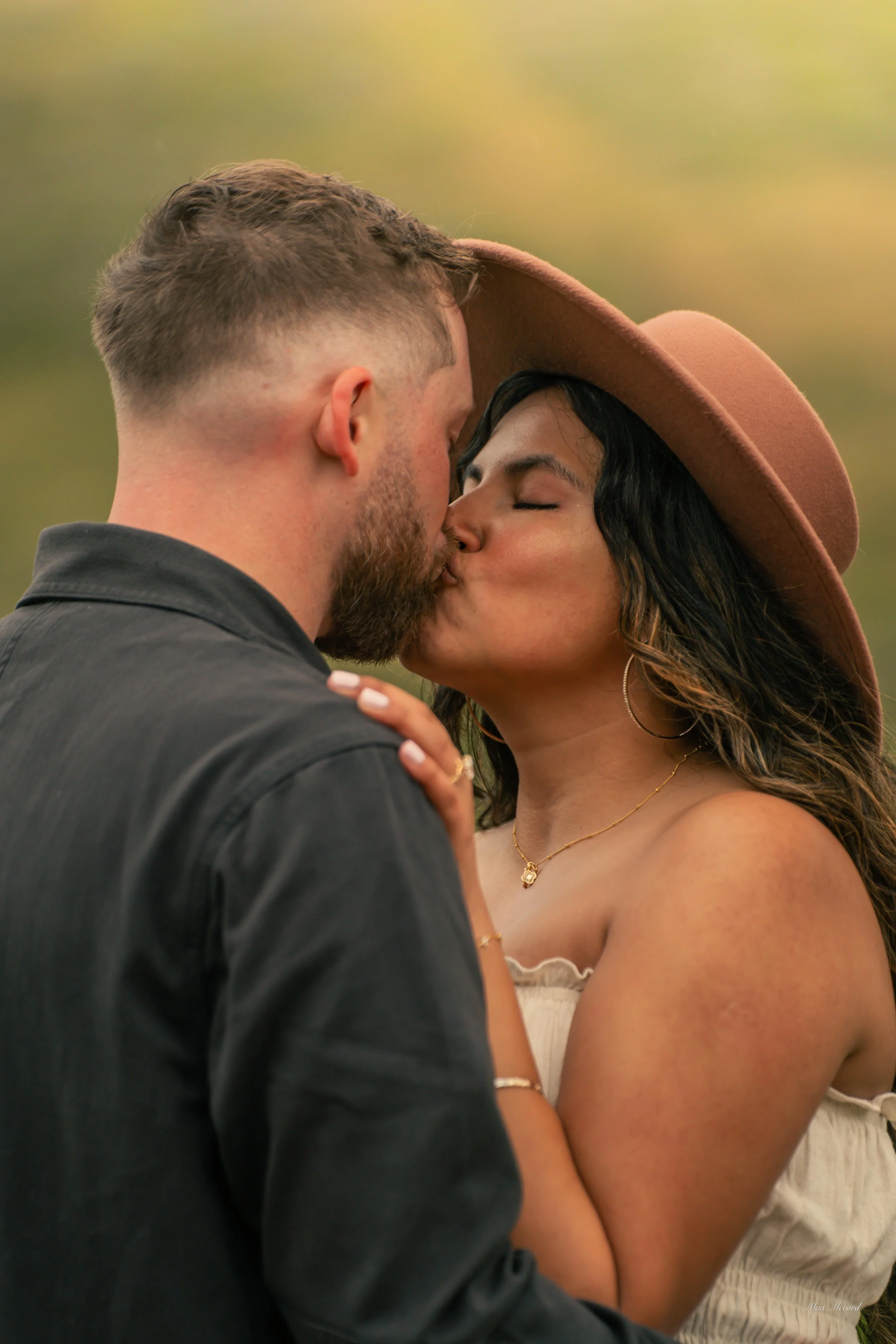 A couple sharing a kiss outdoors, with the woman wearing a large brown hat and gold jewelry, and the man with short hair and a beard, dressed in a black shirt. The background is blurred with warm greens and yellows.