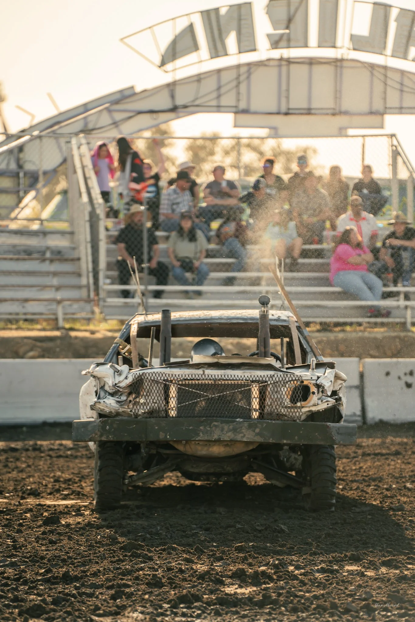 Damaged race car on dirt track with crowd of spectators in bleachers in background.