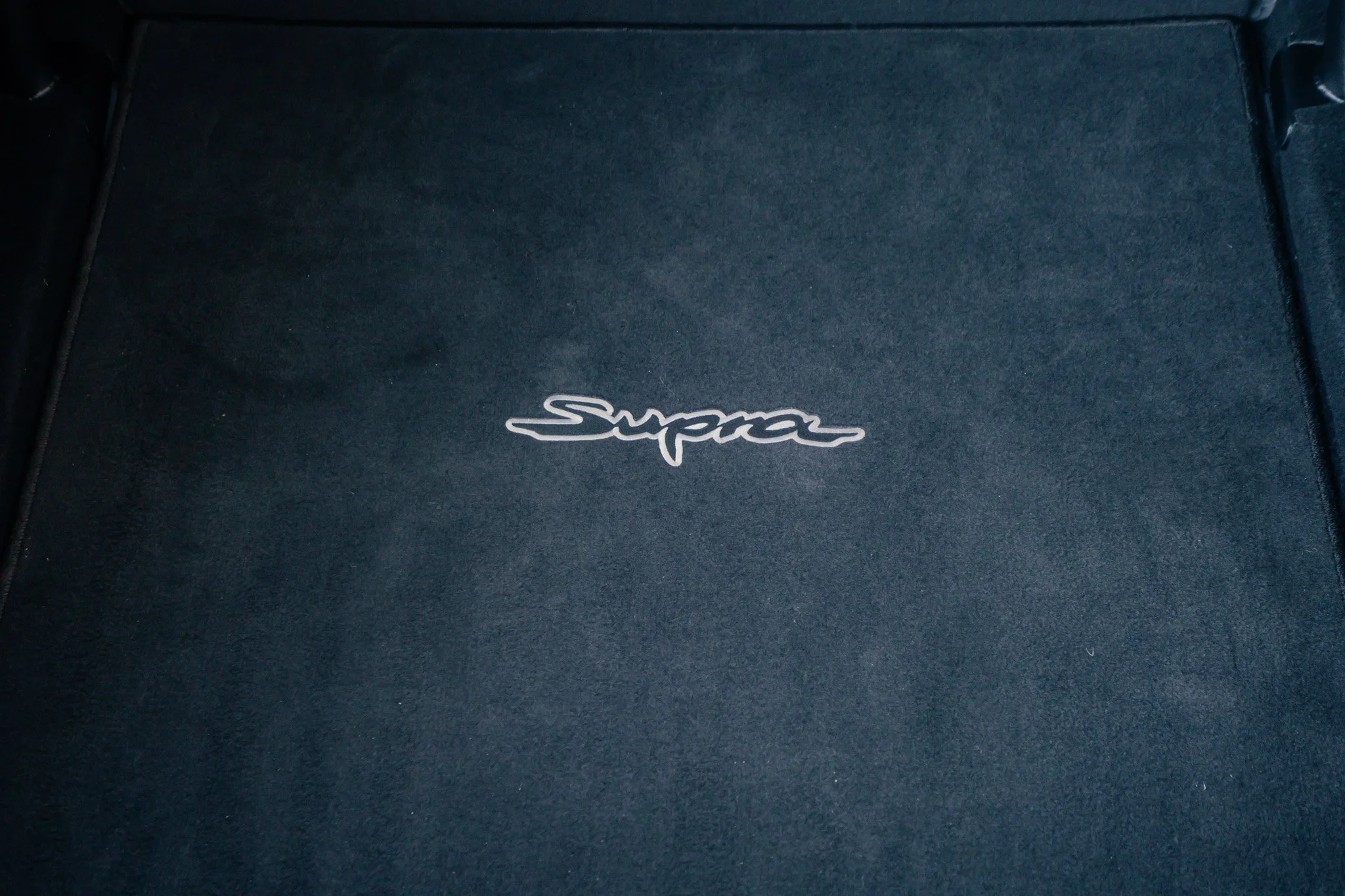 Close-up of a black car floor mat with a silver embroidered 'Supra' logo in the center.
