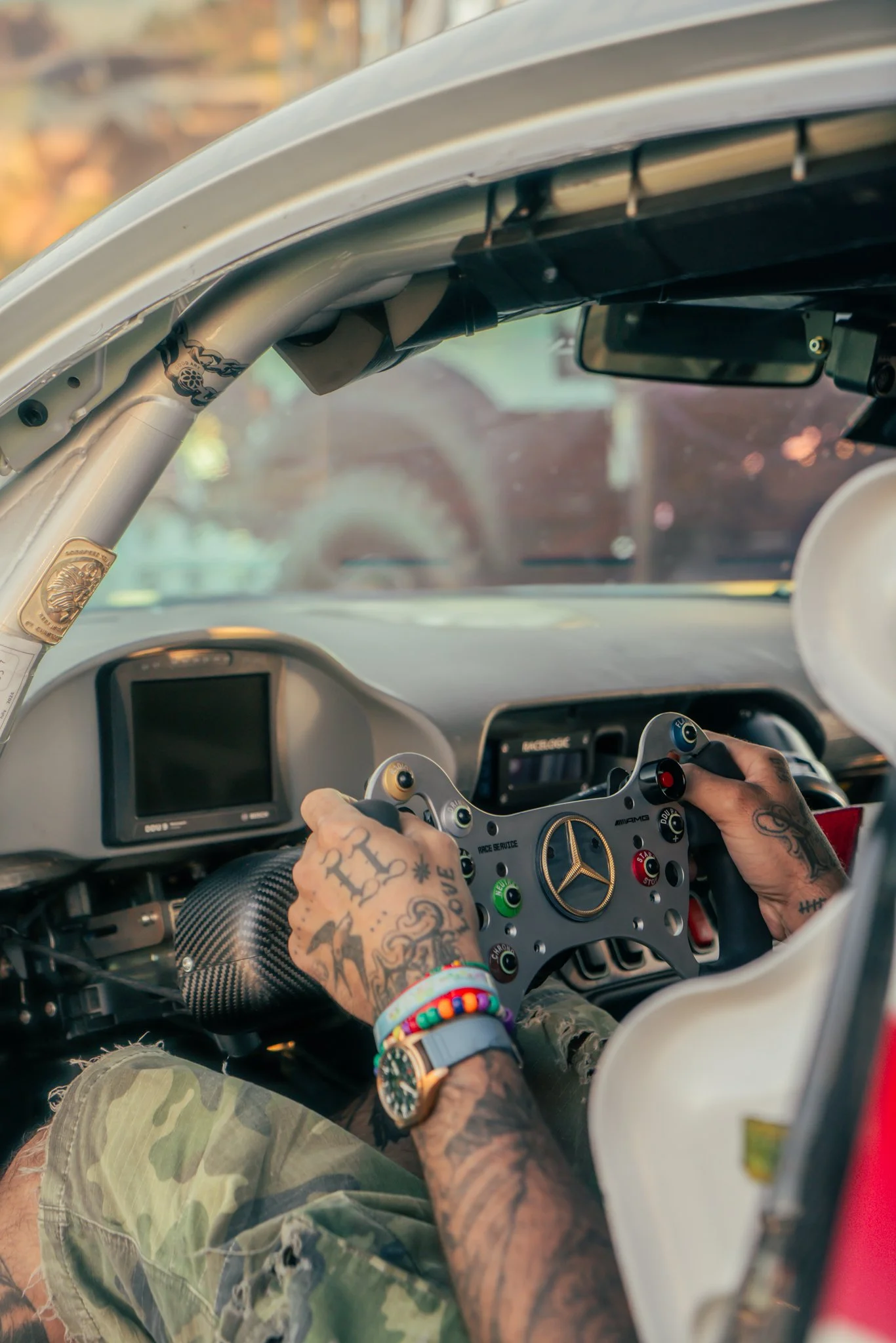 Person with tattoos on hands gripping a race car steering wheel inside a car, wearing colorful bracelets and camouflage shorts.