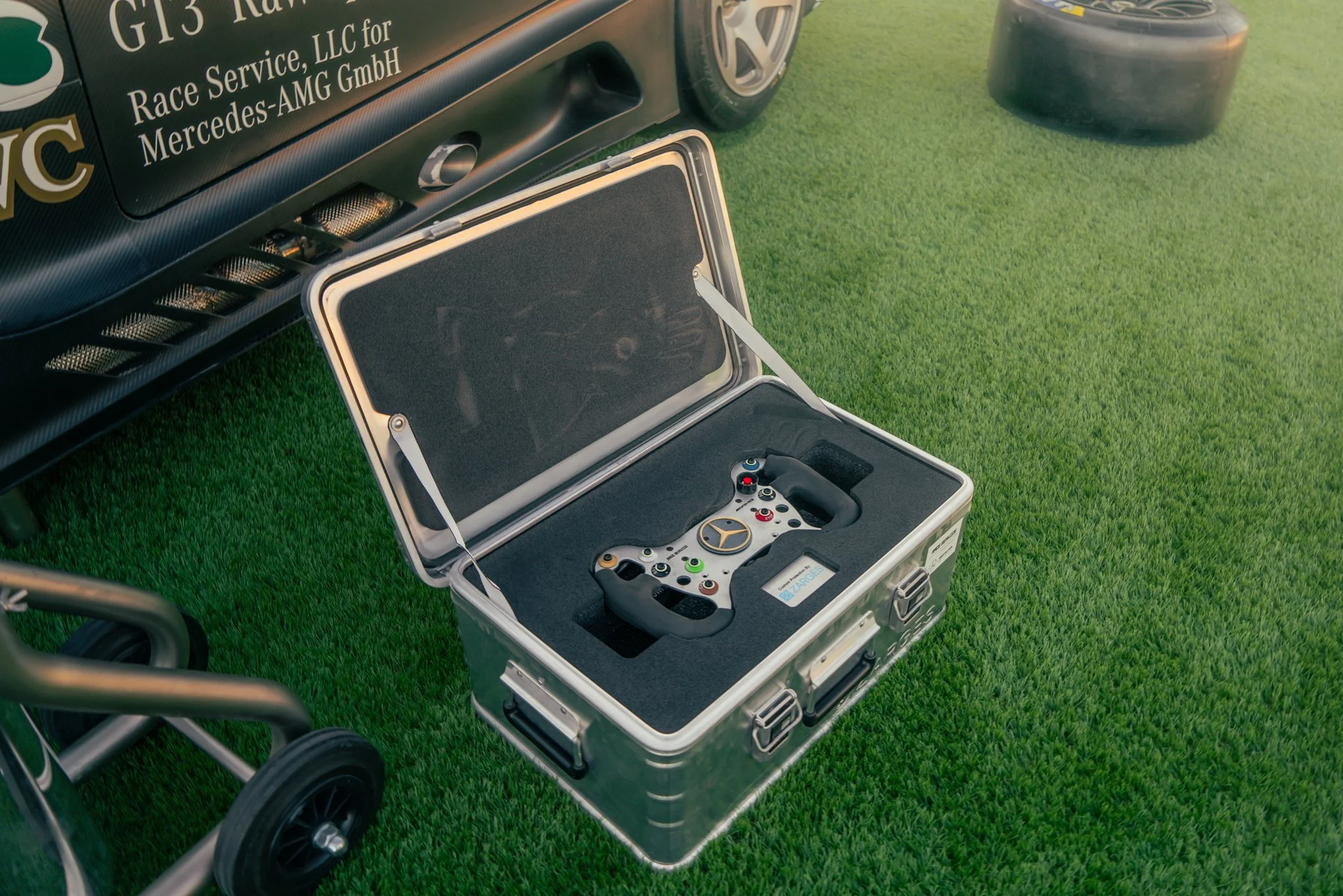 A racing car with branding and a race car steering wheel inside an open protective case on a grassy field.