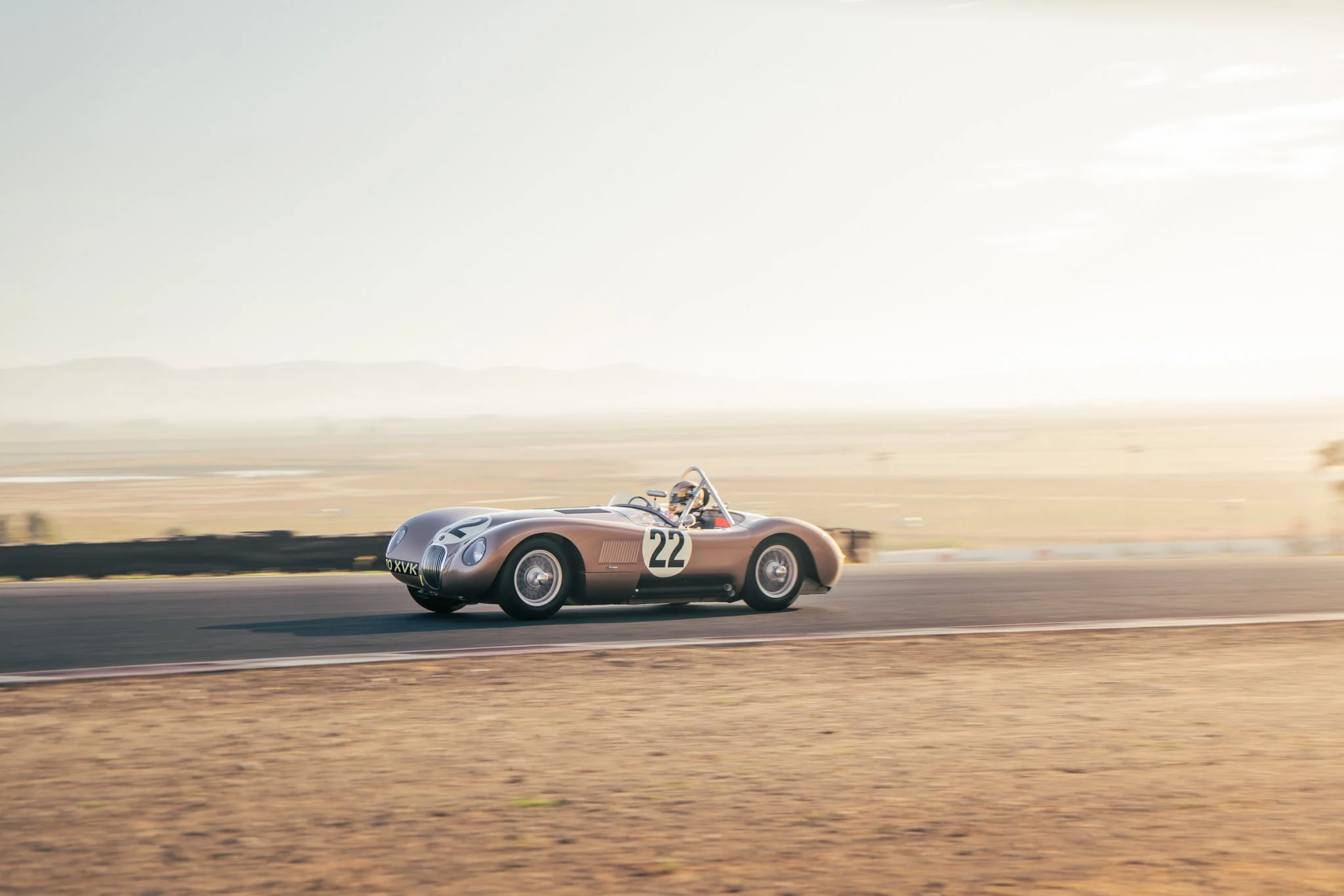 A vintage race car with the number 22 on its side driving on a race track in a desert landscape during sunset.