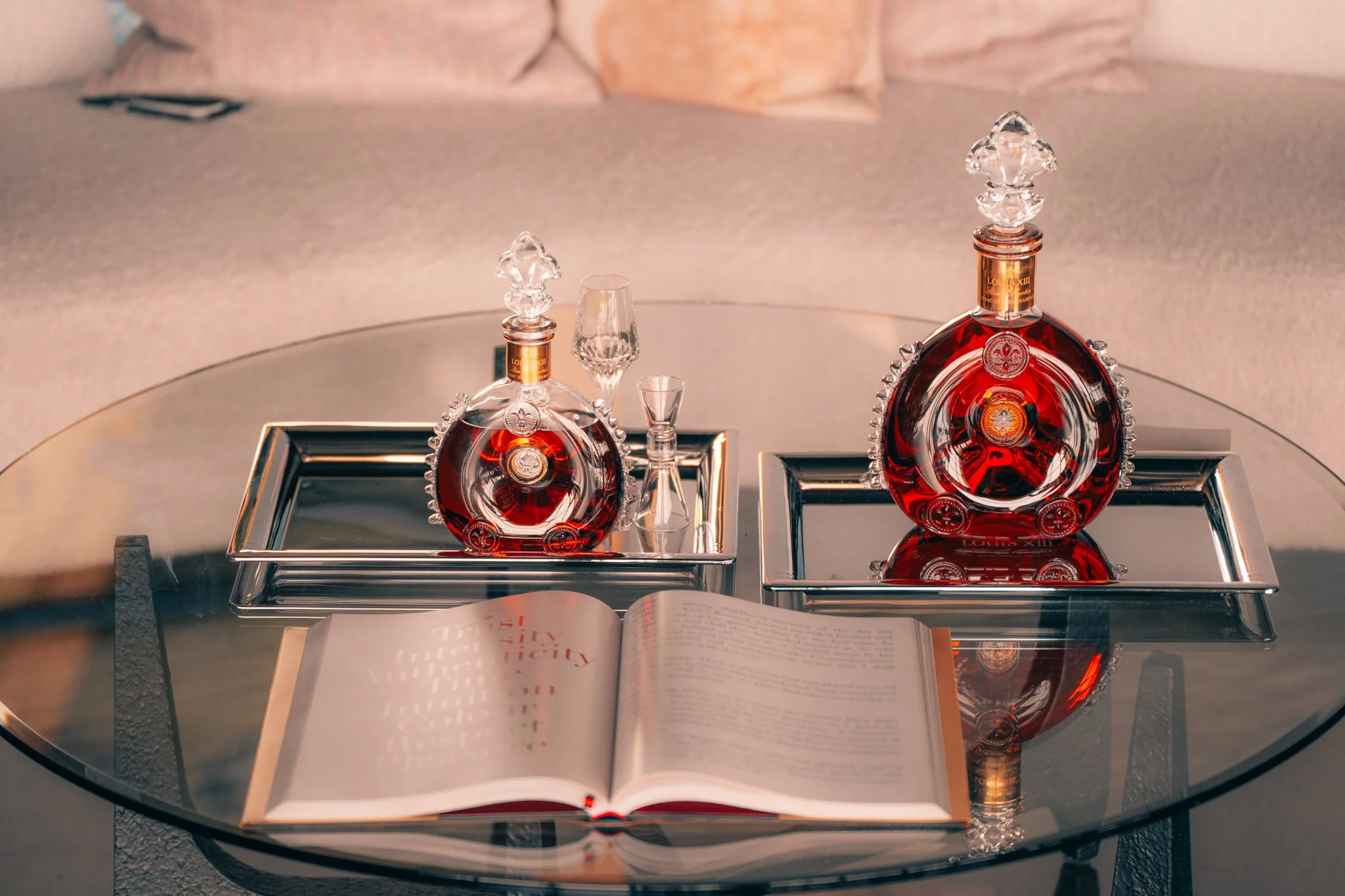 Two decorative bottles of Louis XIII cognac on metal trays, an open book, and a hardcover book on a glass table.