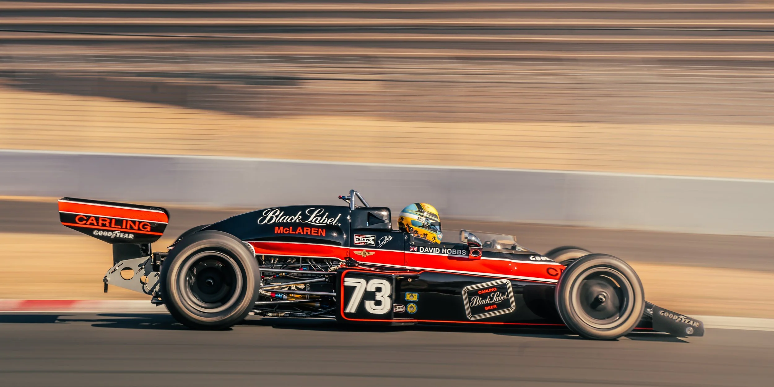 A black and red vintage Formula 1 race car speeding on a track, with the driver wearing a yellow helmet, sponsors' logos including McLaren, Black Label, and Goodyear, and the number 73 on the side.