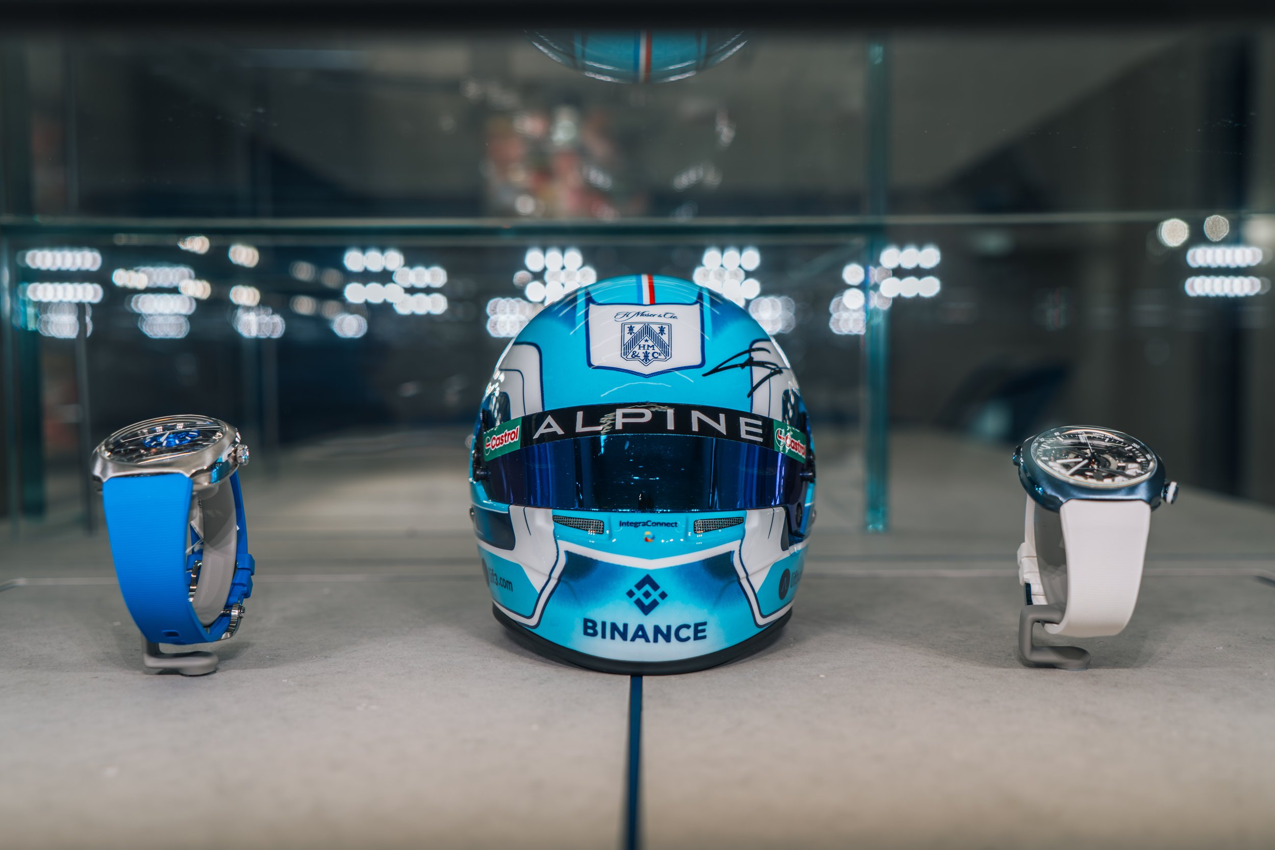 A racing helmet with blue, white, and black colors displayed between two smartwatches on a table, with a glass display case and blurred lights in the background.