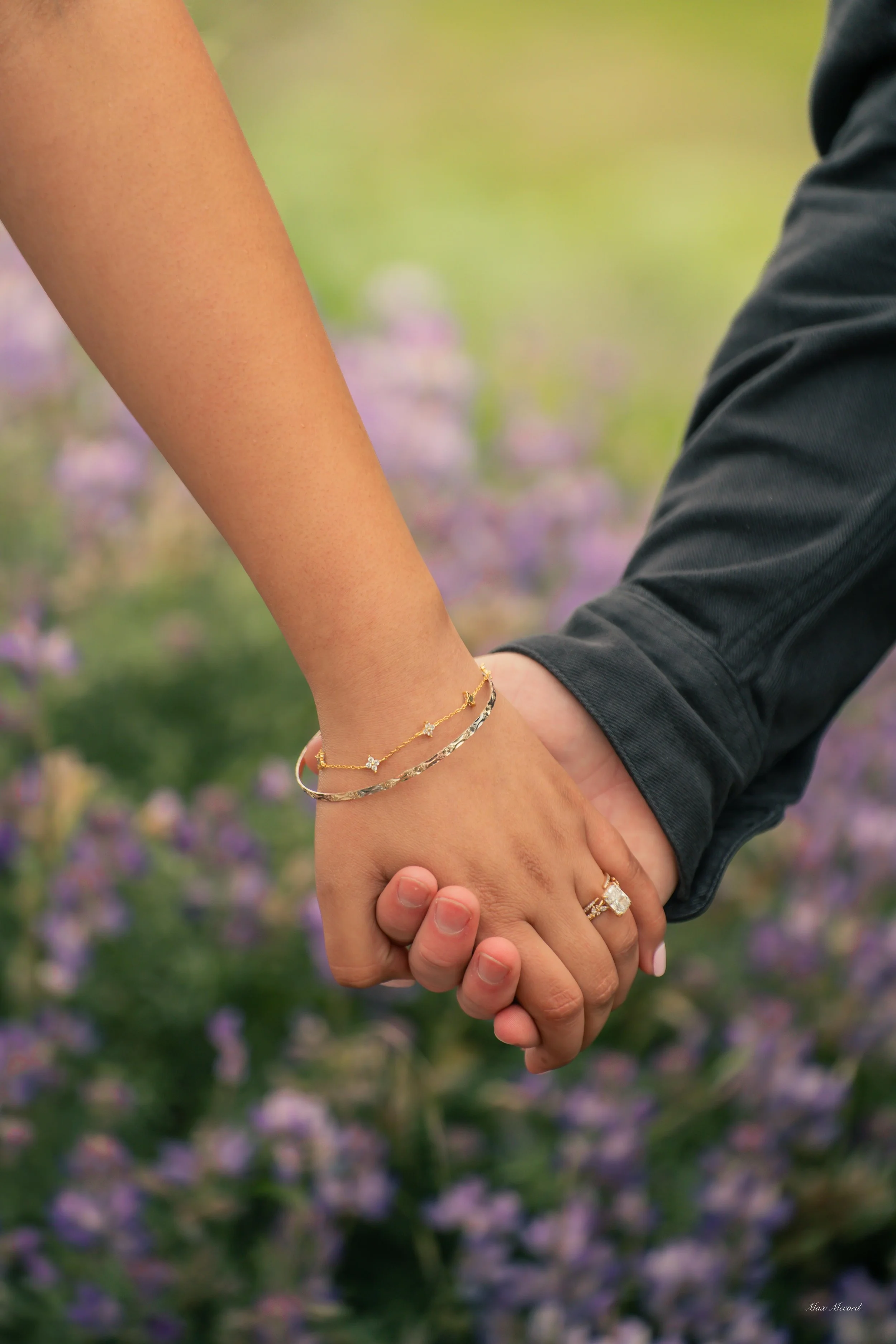 Close-up of couple holding hands, displaying wedding and engagement rings, against a blurred background of purple flowers.
