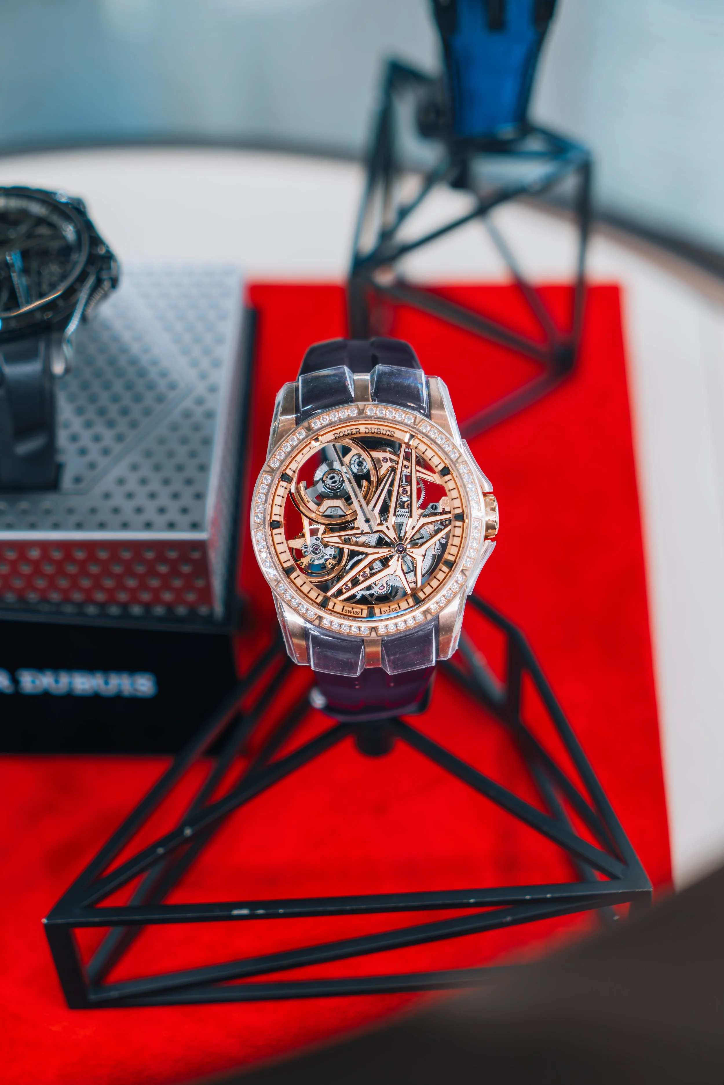 Close-up of a luxury watch with a skeleton design, placed on a red surface with a black geometric stand behind it and another watch to the side.