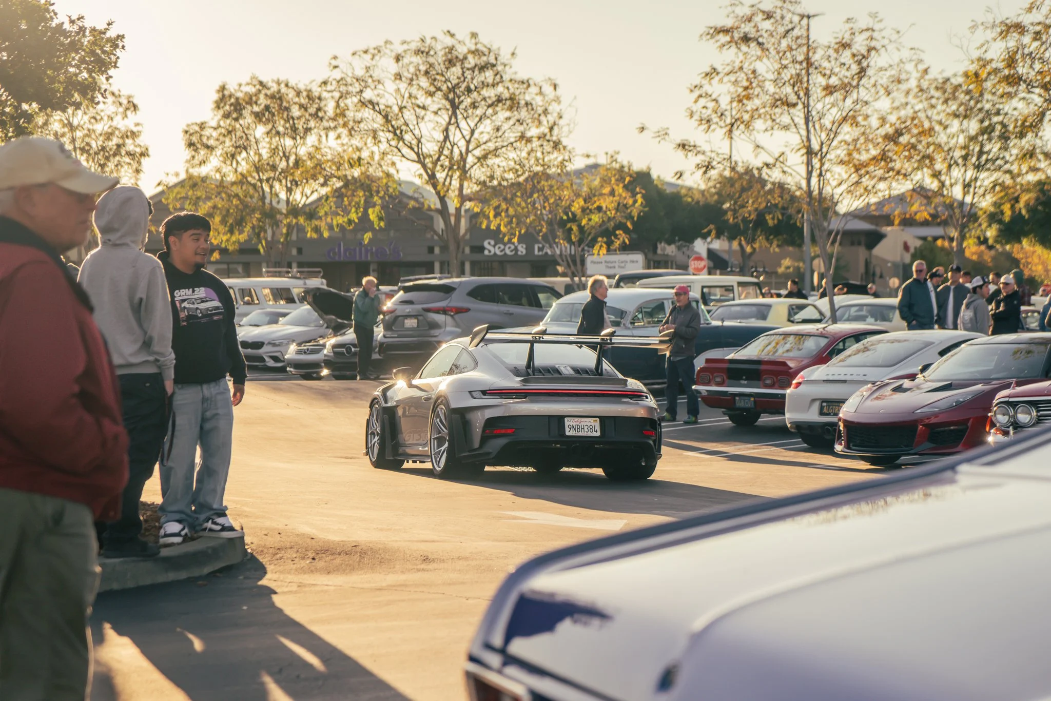 A black sports car with a spoiler driving through a parking lot filled with various cars and people during sunset.