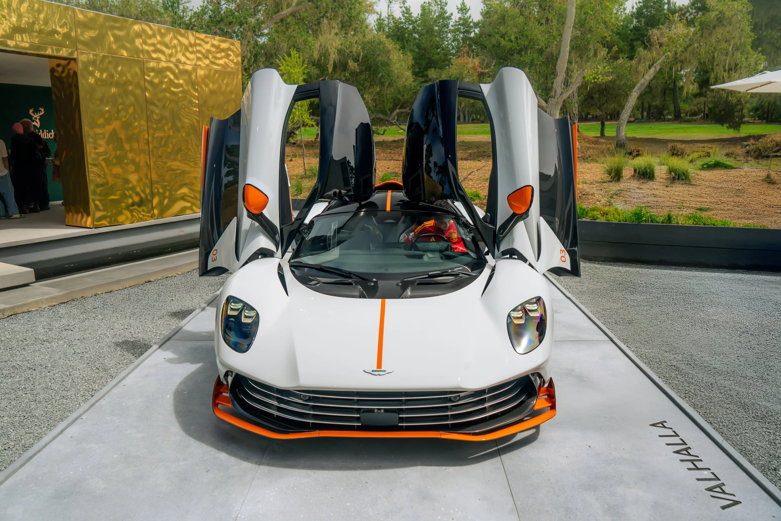 White Pagani Huayra with black and orange accents, gullwing doors open, parked on a display platform labeled 'Pagani Huayra' outdoors with trees and a modern gold building in the background.