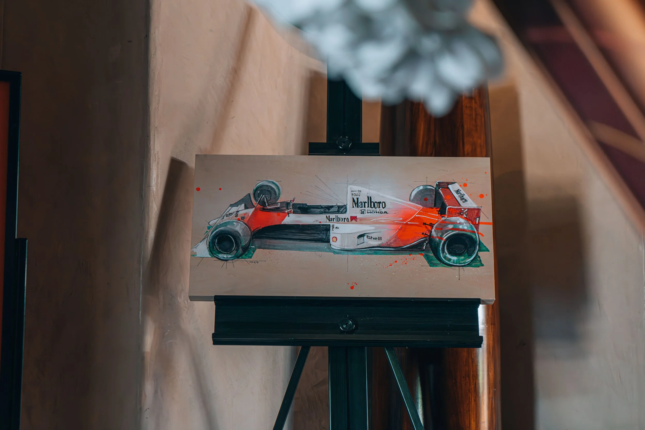 A painting of a vintage Formula 1 race car on an easel in a room with wooden walls.