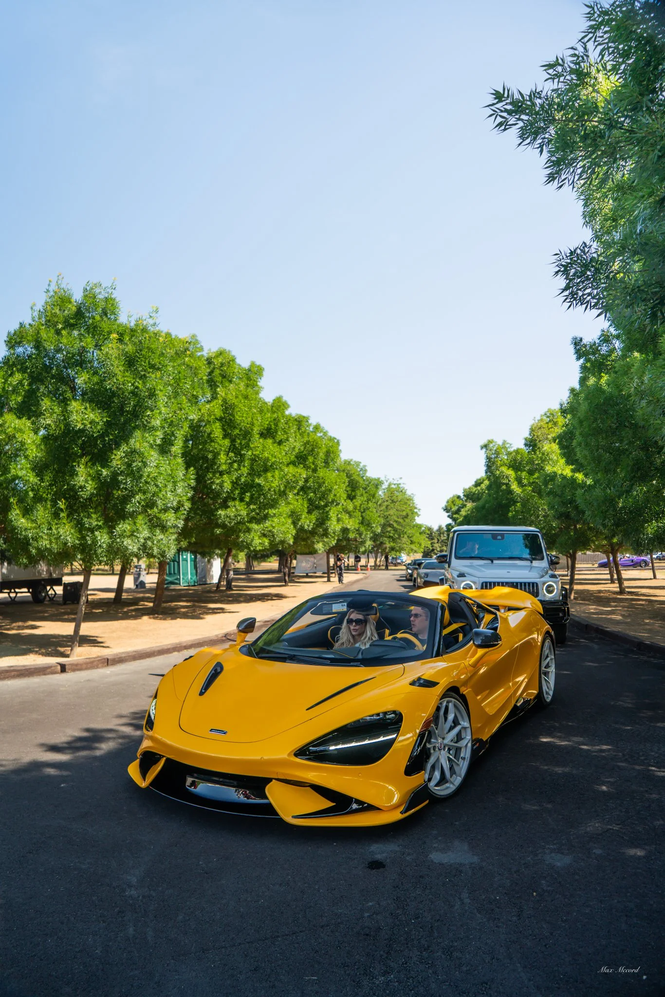 A yellow sports car with two people inside, driving on a paved road lined with green trees. A black SUV is behind the sports car, and there are other vehicles in the background under a clear blue sky.