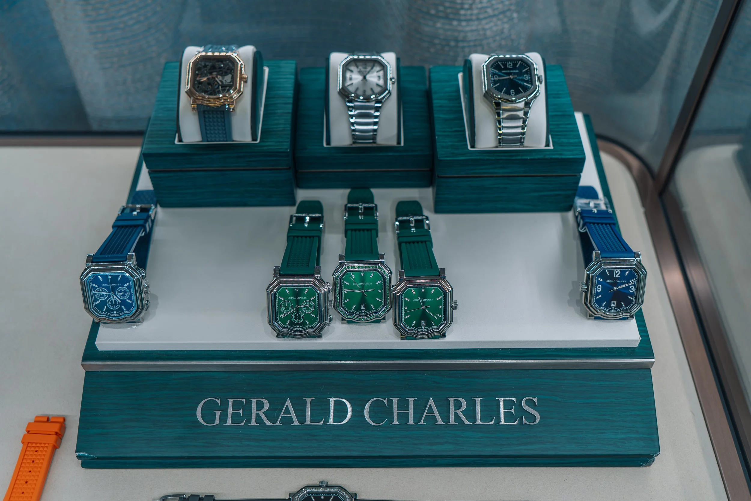 Display of luxury watches and watch straps on a table with a Gerad Charles sign. The watches are in a case, and some are on green and blue straps. The image showcases a collection of high-end watches with various designs and styles.