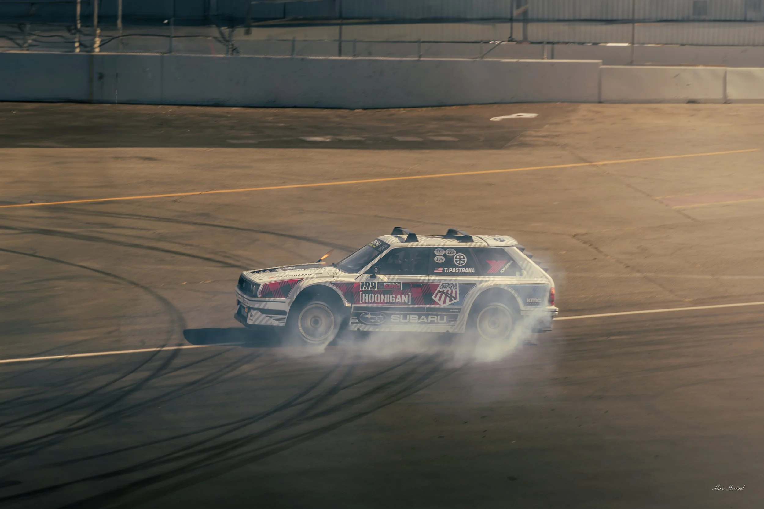 A race car drifting on a race track, producing smoke from its tires, with tire marks on the asphalt and a concrete barrier in the background.
