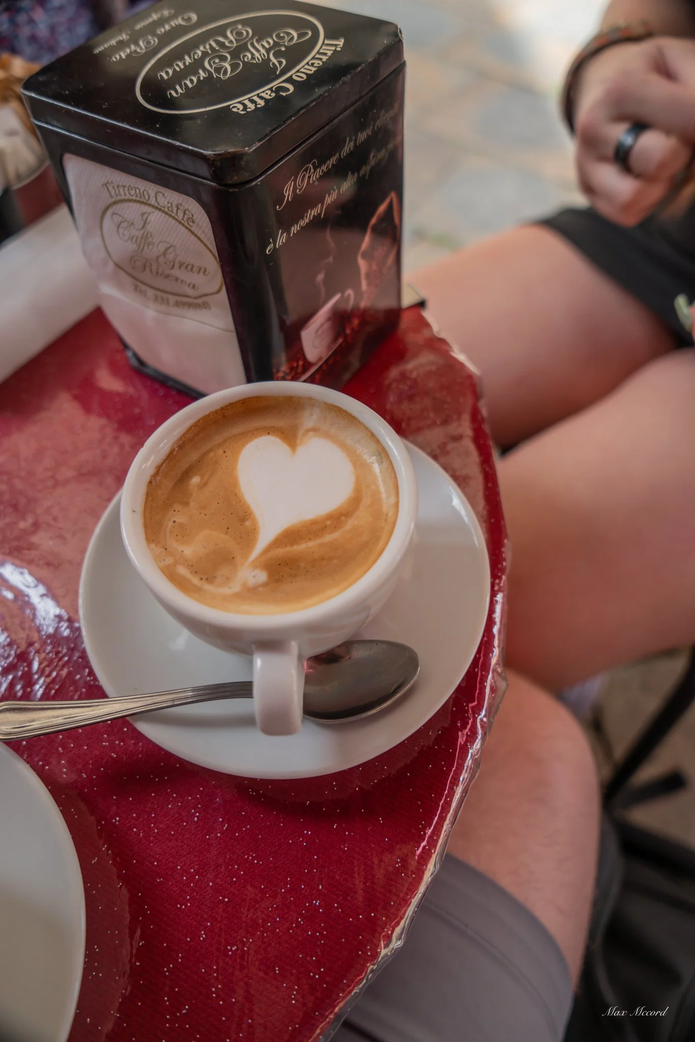 A small white cup of cappuccino with heart-shaped latte art on top, resting on a matching saucer with a spoon, placed on a shiny red tablecloth. Behind it is a black box of Tirreno Caffe. To the right, a person's arm and knee are partially visible.