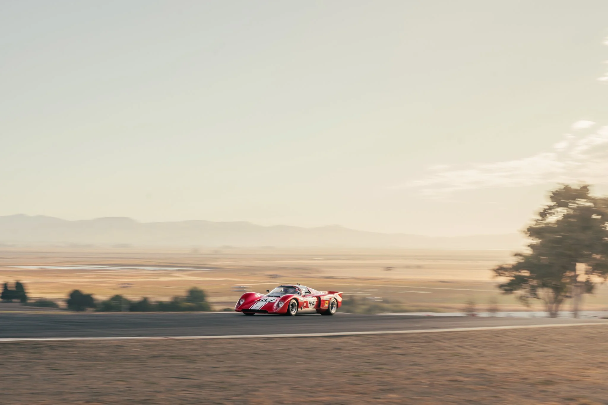 A vintage red racing car with a white stripe on the track in a desert landscape with mountains in the background and a few trees.