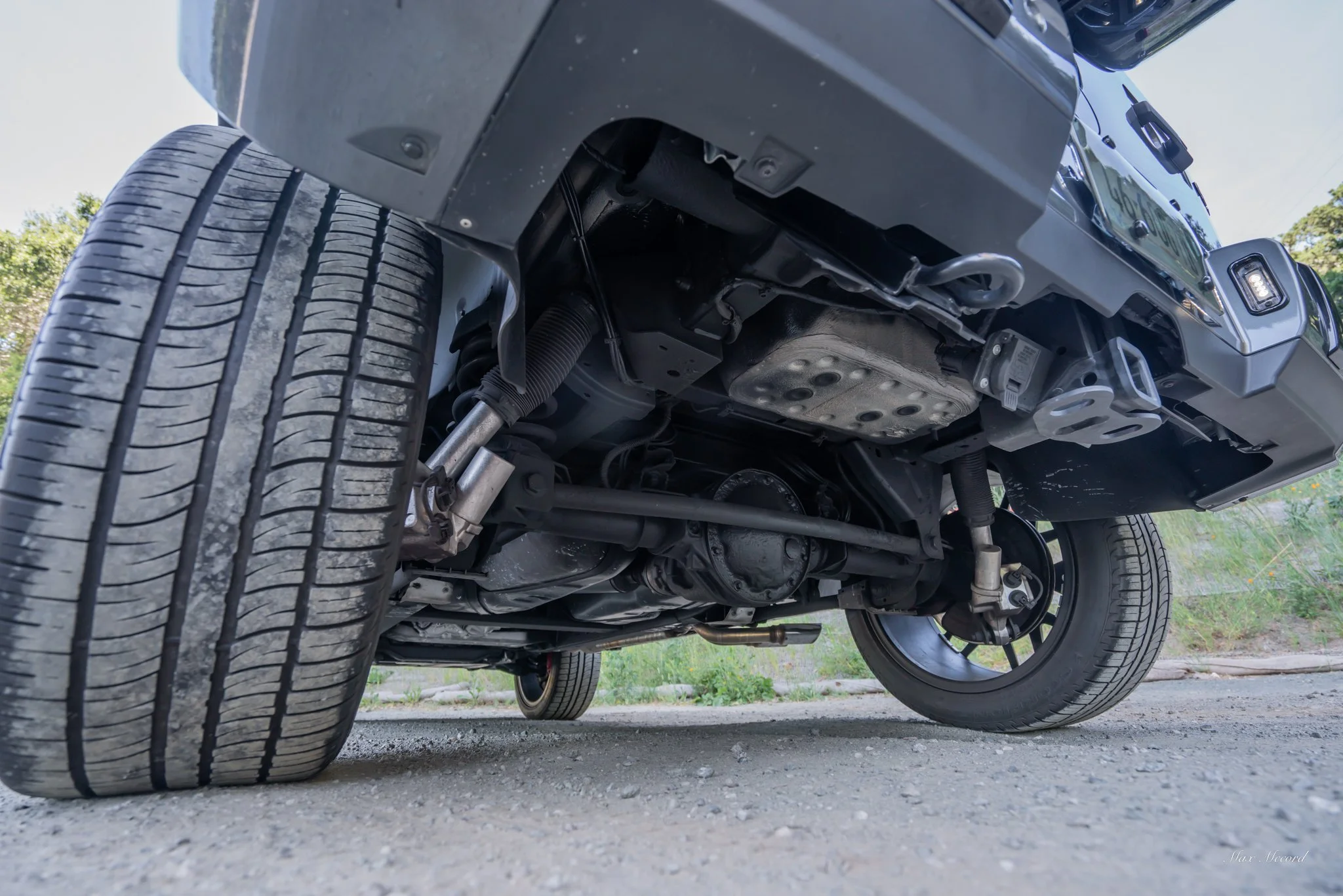 Underneath view of a vehicle showing the suspension system, differential, and tires on a gravel surface with greenery in the background.