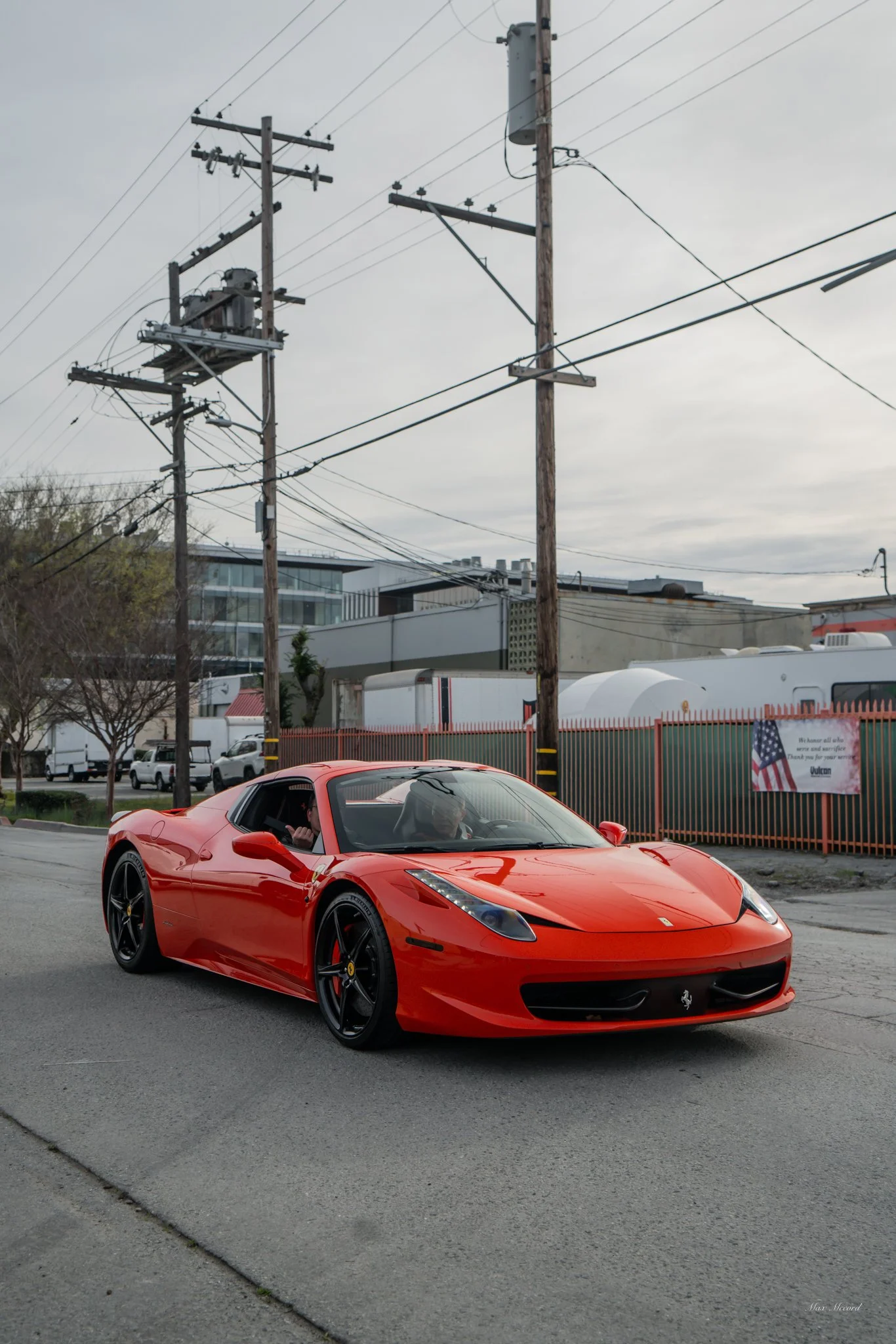 Red Ferrari sports car driving on a city street with power lines and industrial buildings in the background.