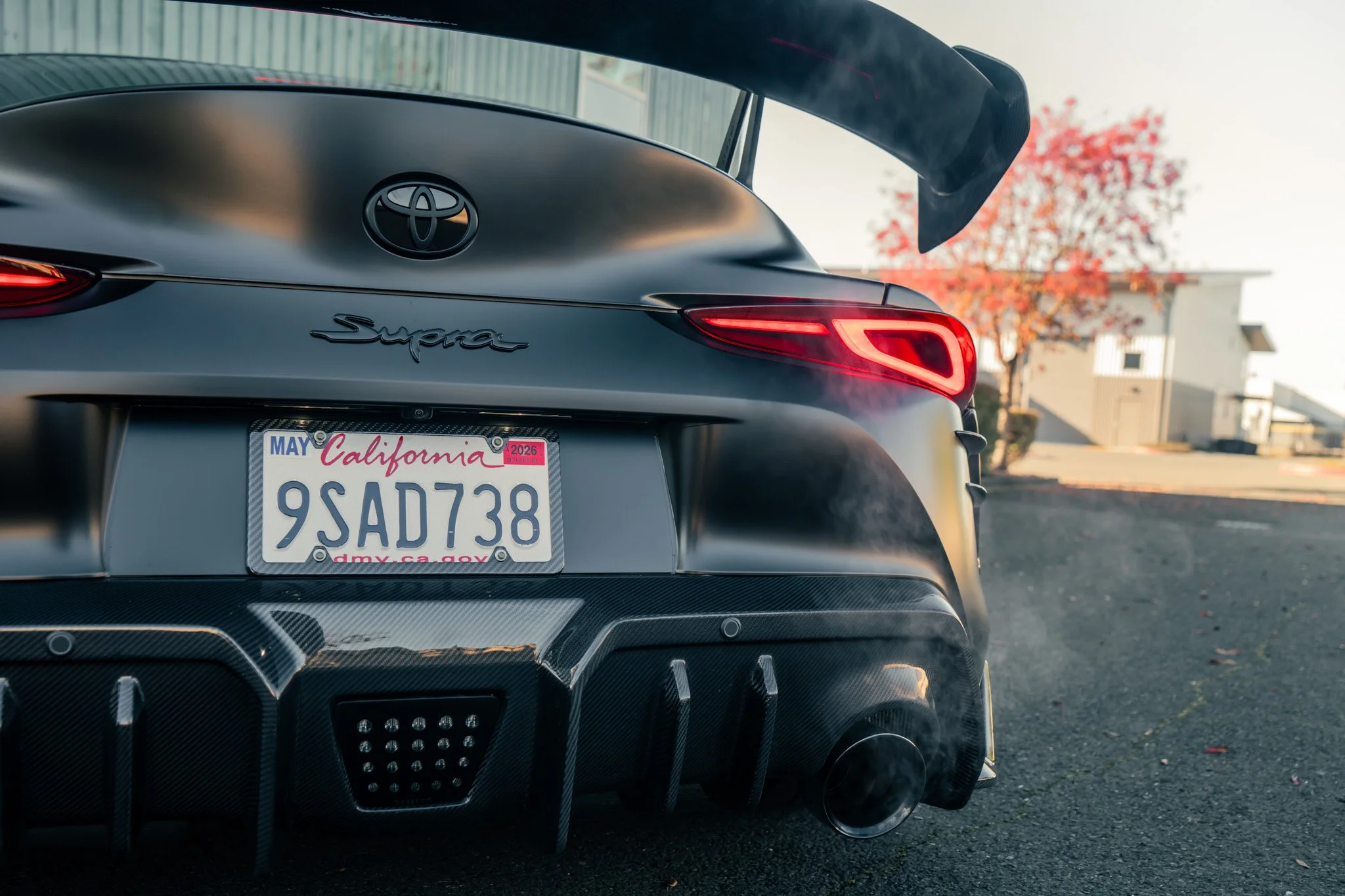 Rear view of a matte black Toyota Supra with a custom exhaust, showing smoke emitting from the exhaust pipe, parked on a driveway with a house and autumn-colored tree in the background.