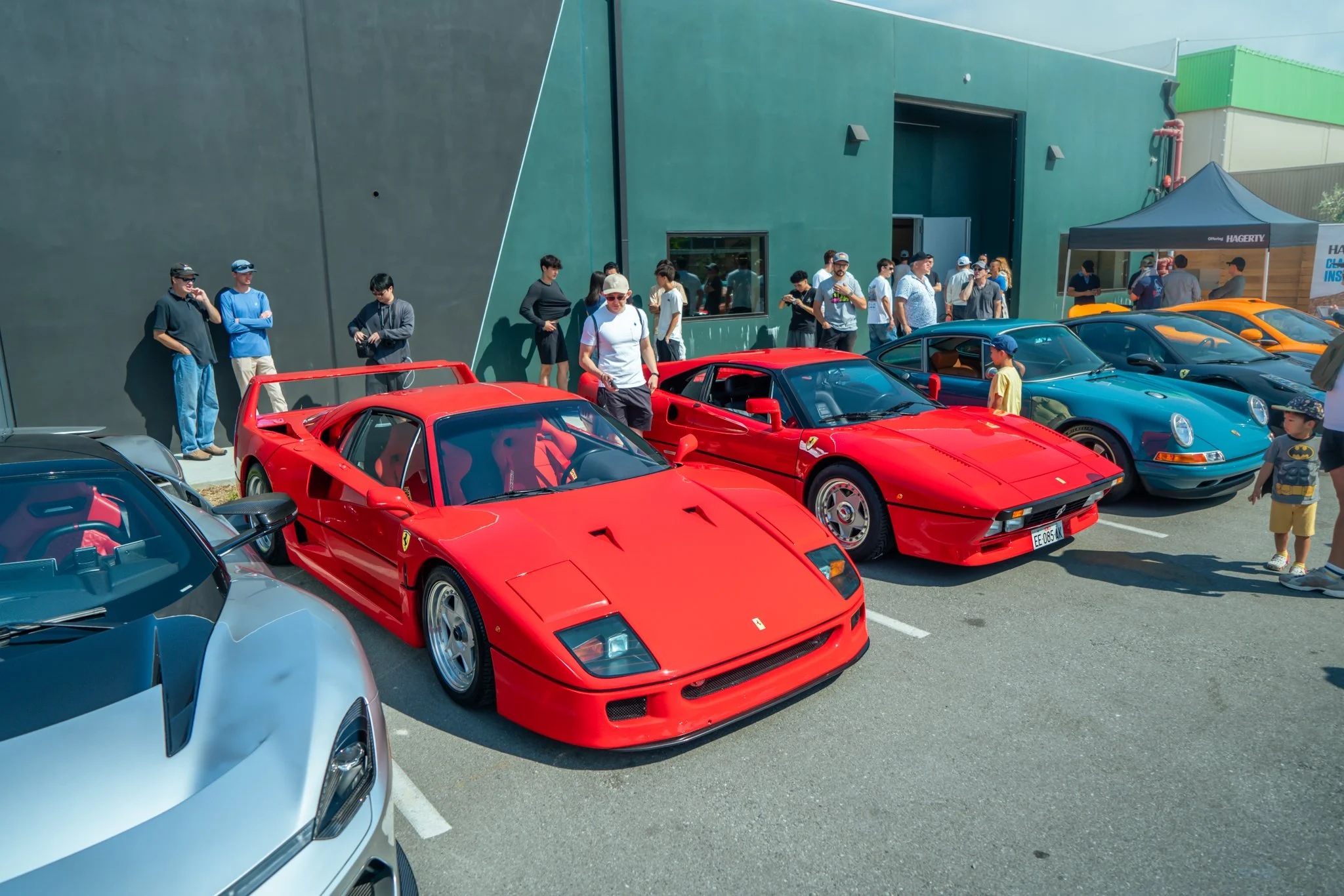 A row of red and blue classic sports cars, including Ferraris and Porsches, parked outside a modern building during a car show with people walking around and observing the cars.