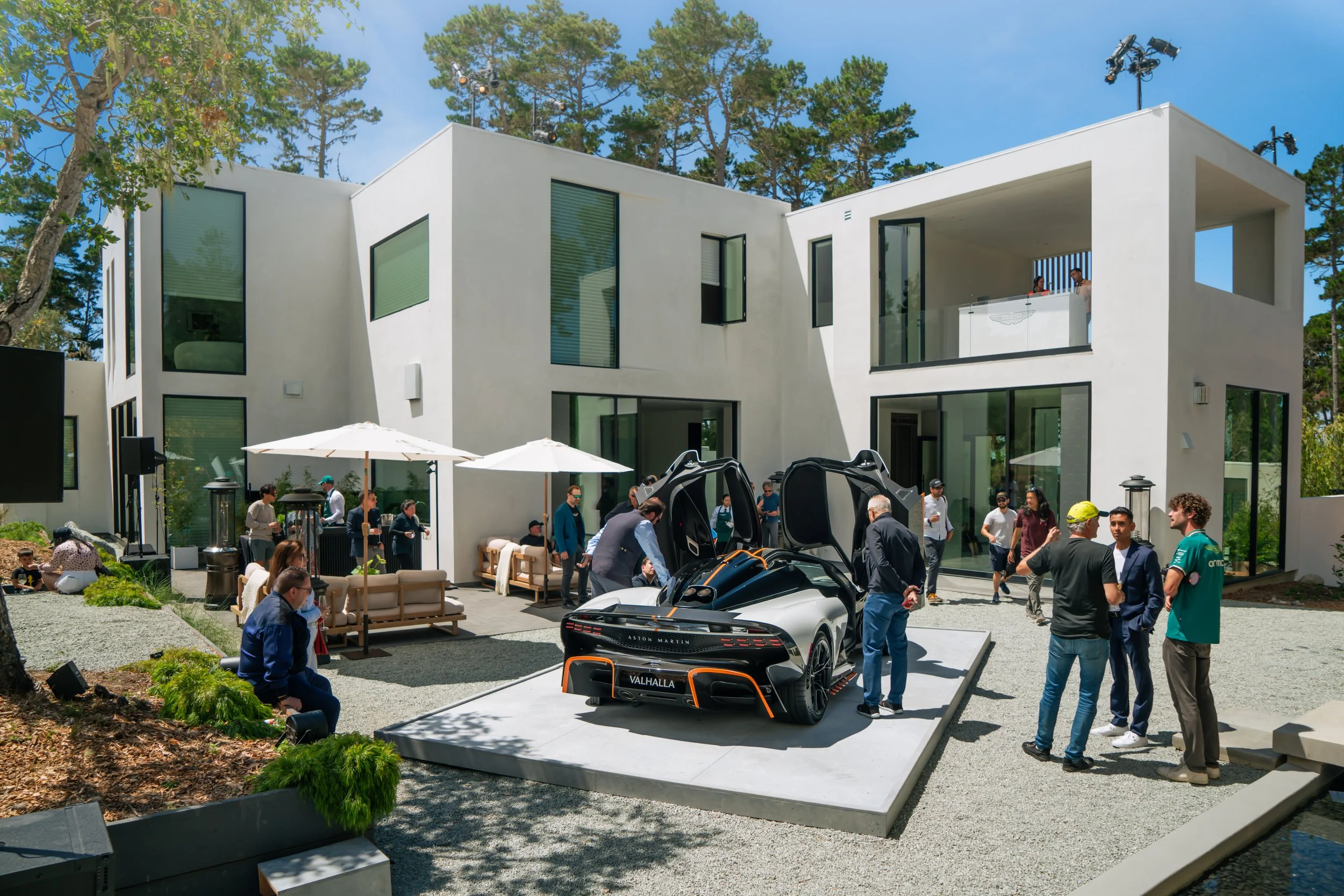 An outdoor event with a white building in the background. A silver sports car with scissor doors open is displayed on a platform in the foreground. People are gathered around, some talking, some looking at the car, under umbrellas and in seats, on a 