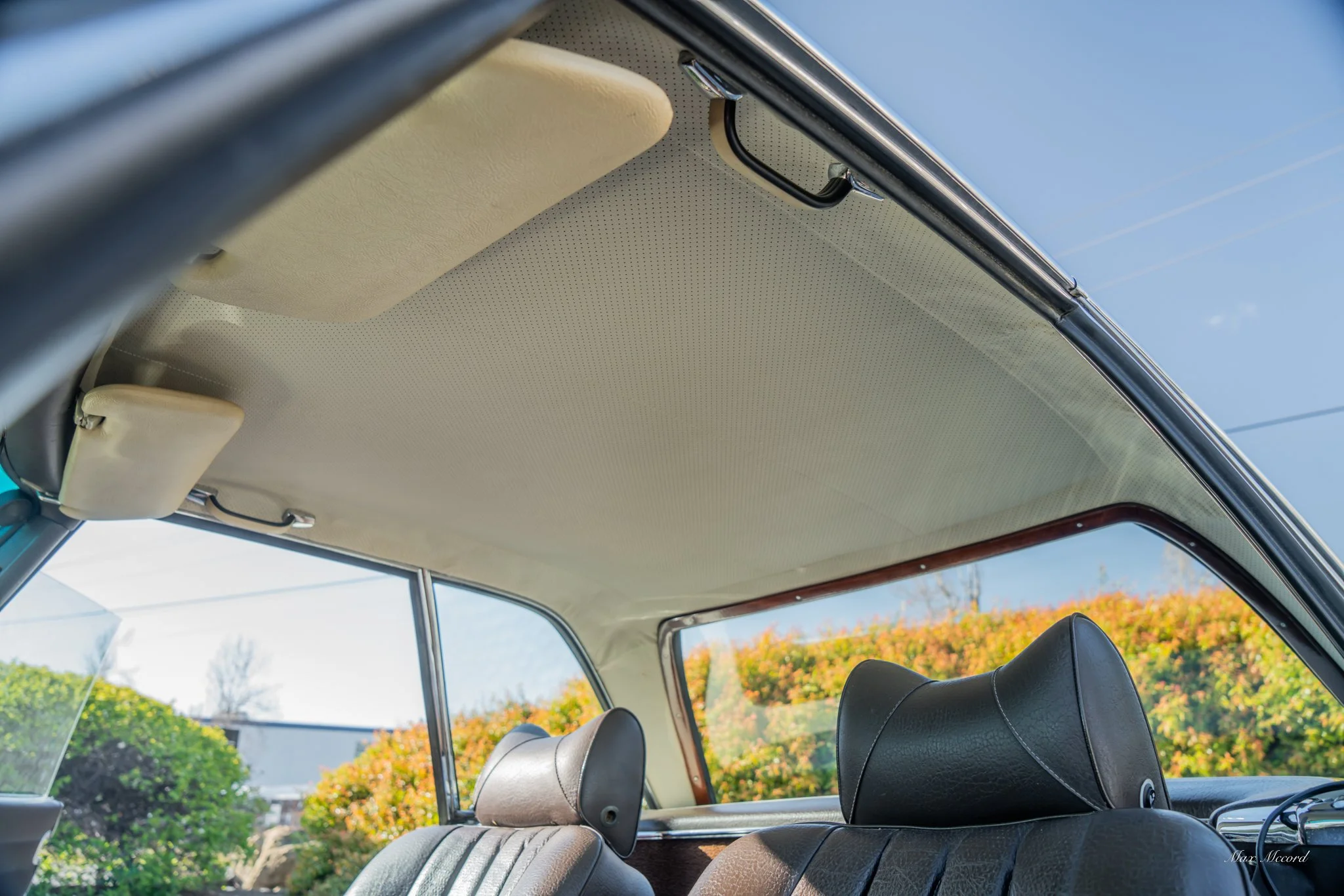 The interior of a vintage car showing the beige headliner, black leather seats, and rear side window with a view of greenery outside.