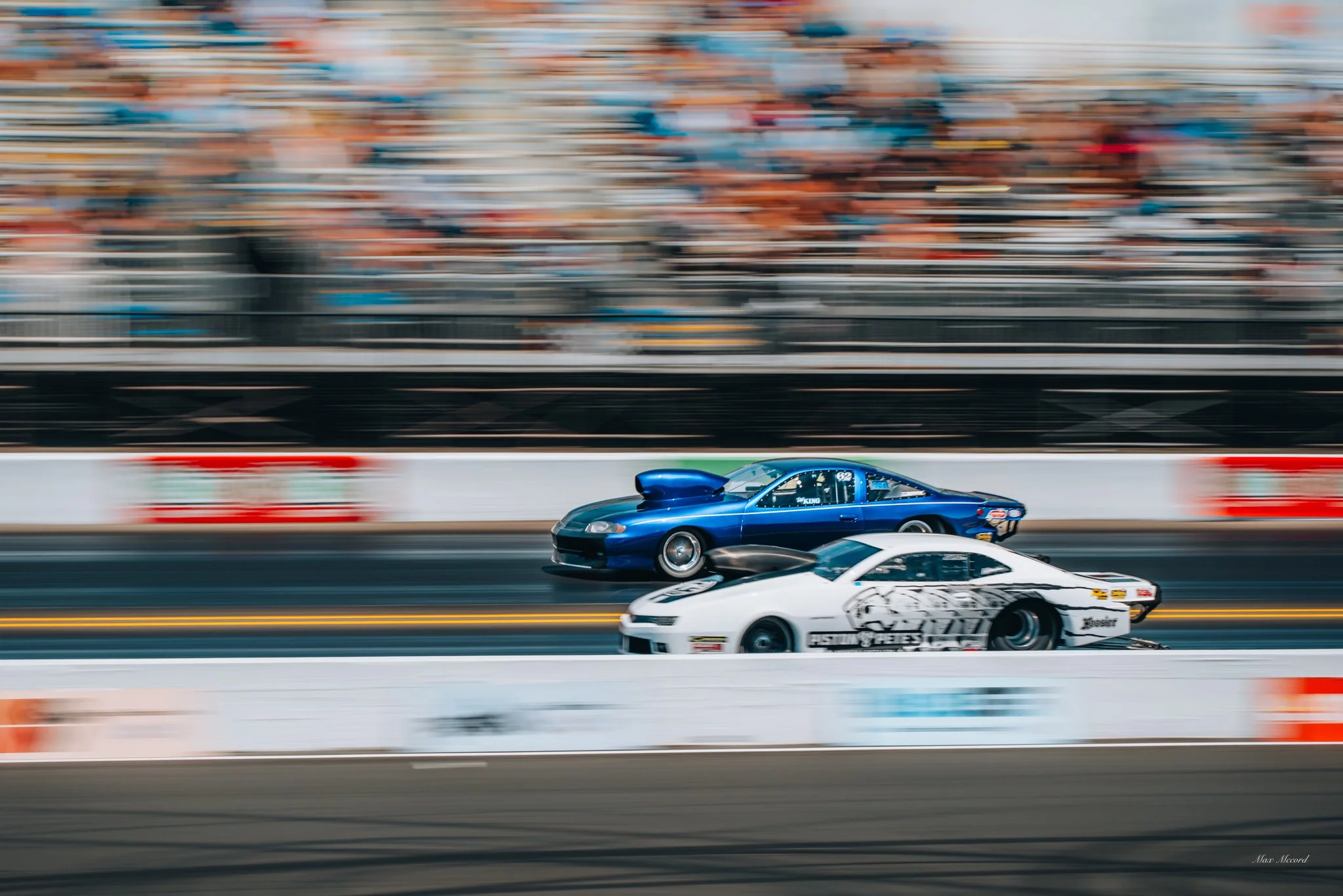 Two drag race cars on the track, one white with black and gray graphics, and one blue with a large hood scoop, racing at a drag strip while blurred spectators watch from the stands.