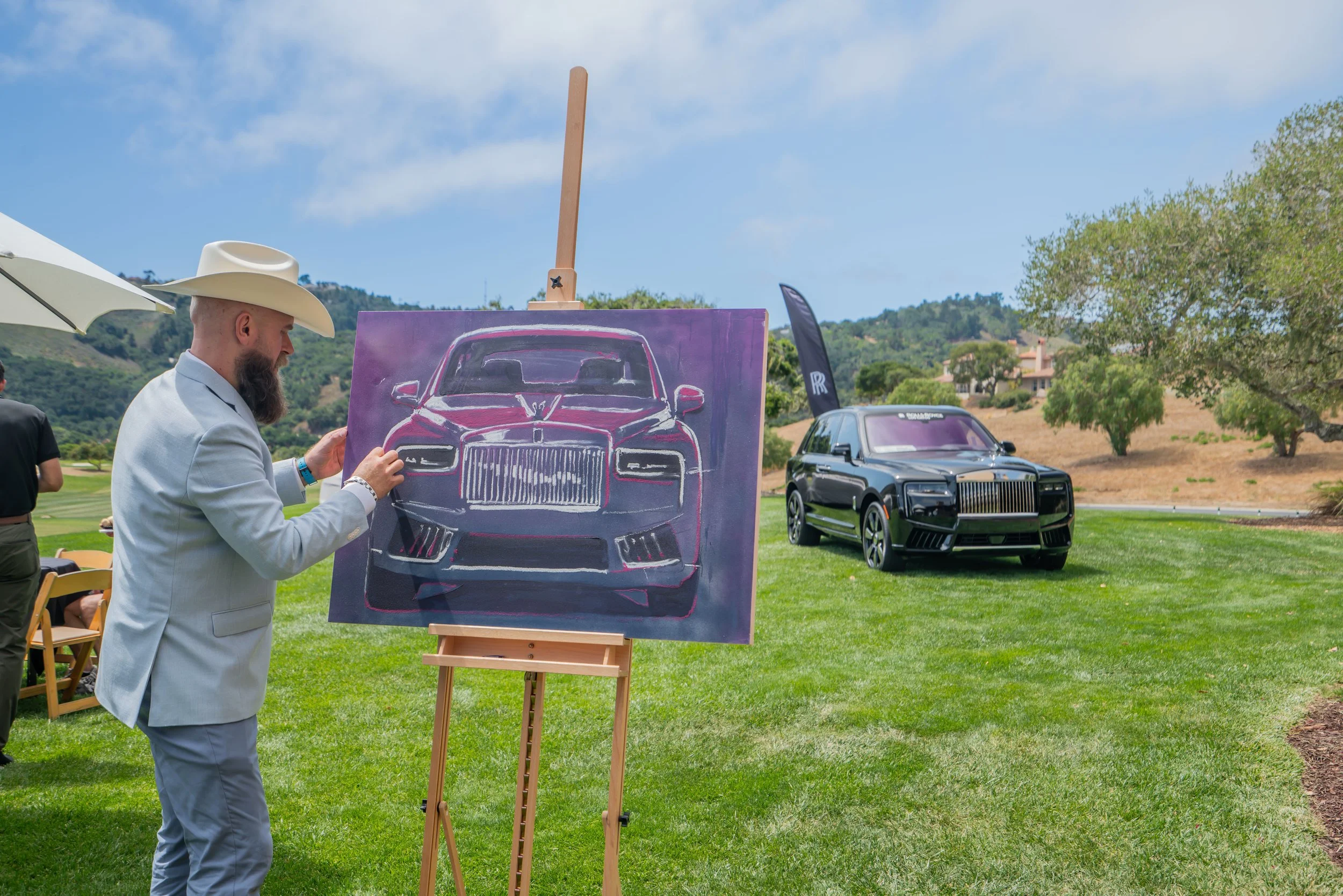 Man with a cowboy hat and suit painting a portrait of a black Rolls-Royce car on canvas outdoors, with a second black Rolls-Royce car parked on the grass nearby, under a partly cloudy sky.