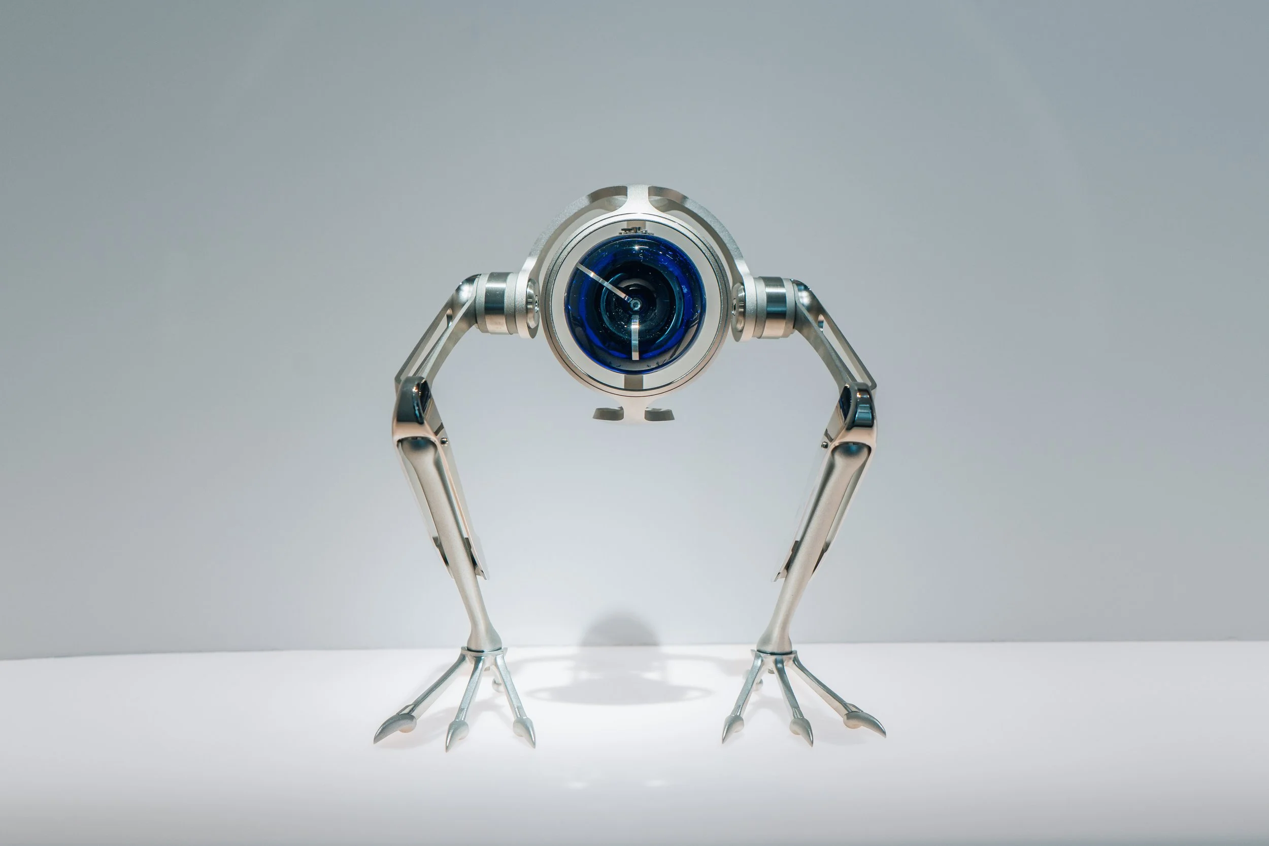 A futuristic robot with a spherical blue camera eye, four metallic limbs, and bird-like feet, standing on a white surface against a plain light gray background.