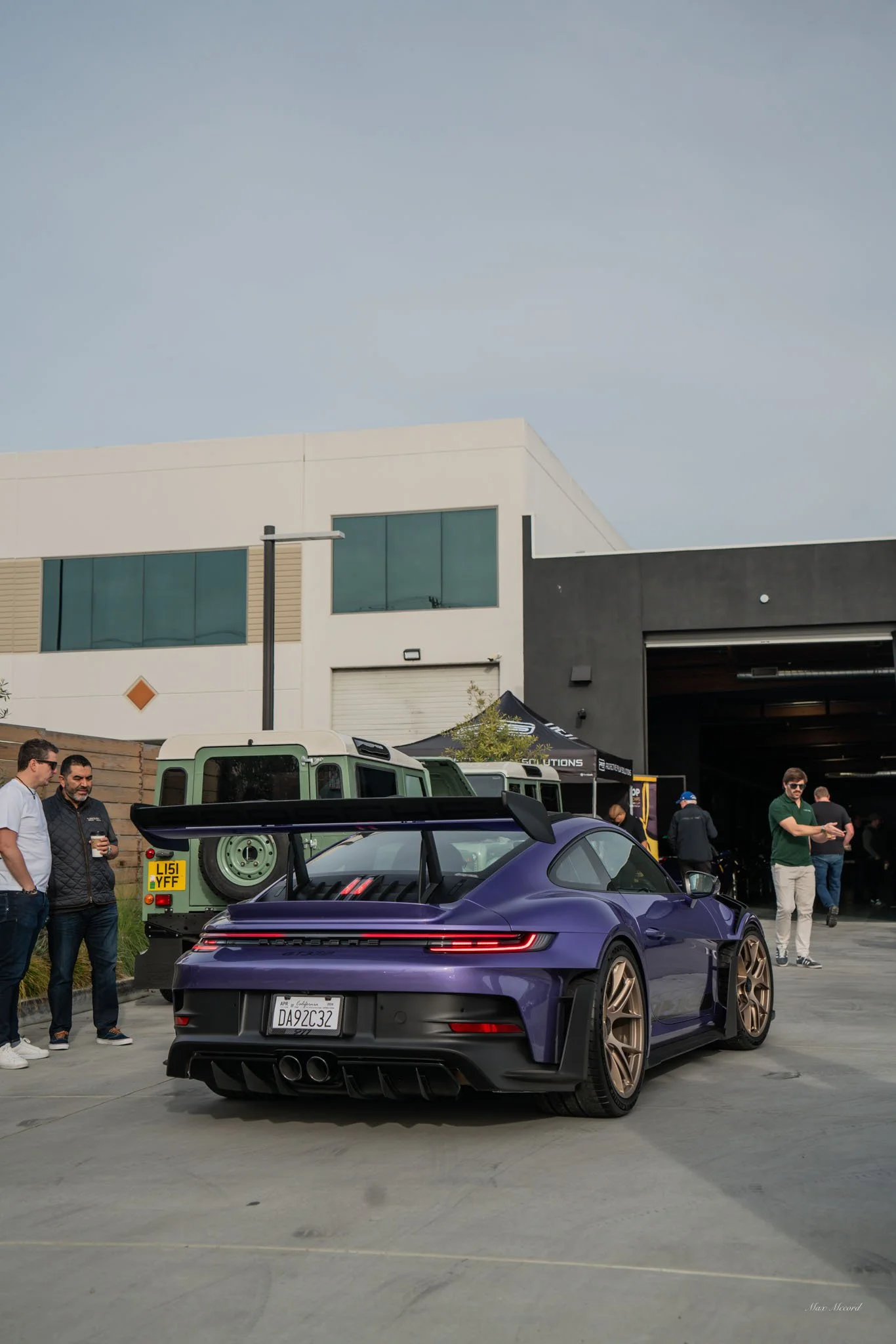 A purple Porsche 911 sports car with a large rear wing parked outdoors at a car event, with several people standing nearby.