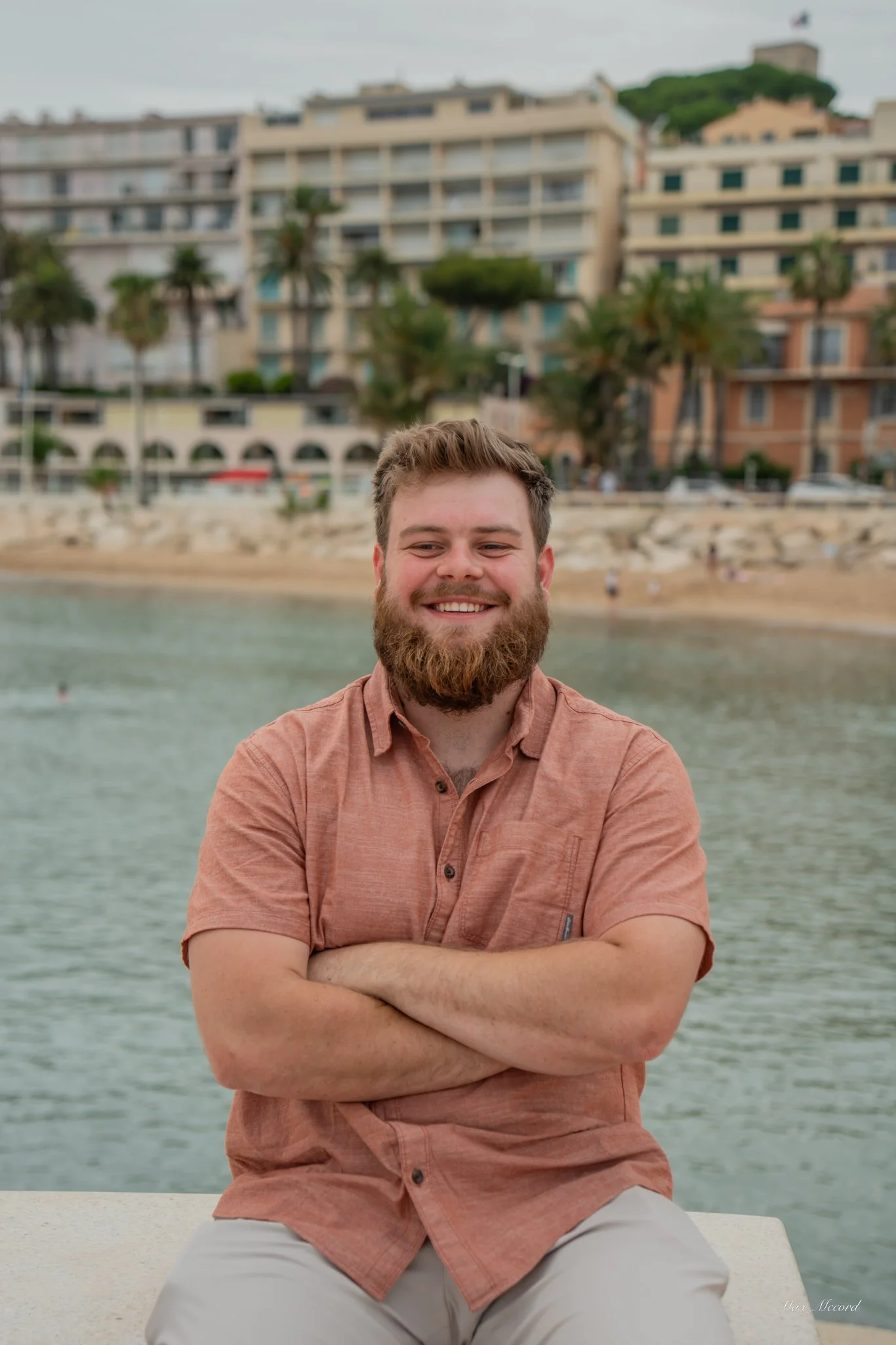 A man with a beard and short hair smiling with crossed arms sits in front of a body of water, with buildings and palm trees in the background.