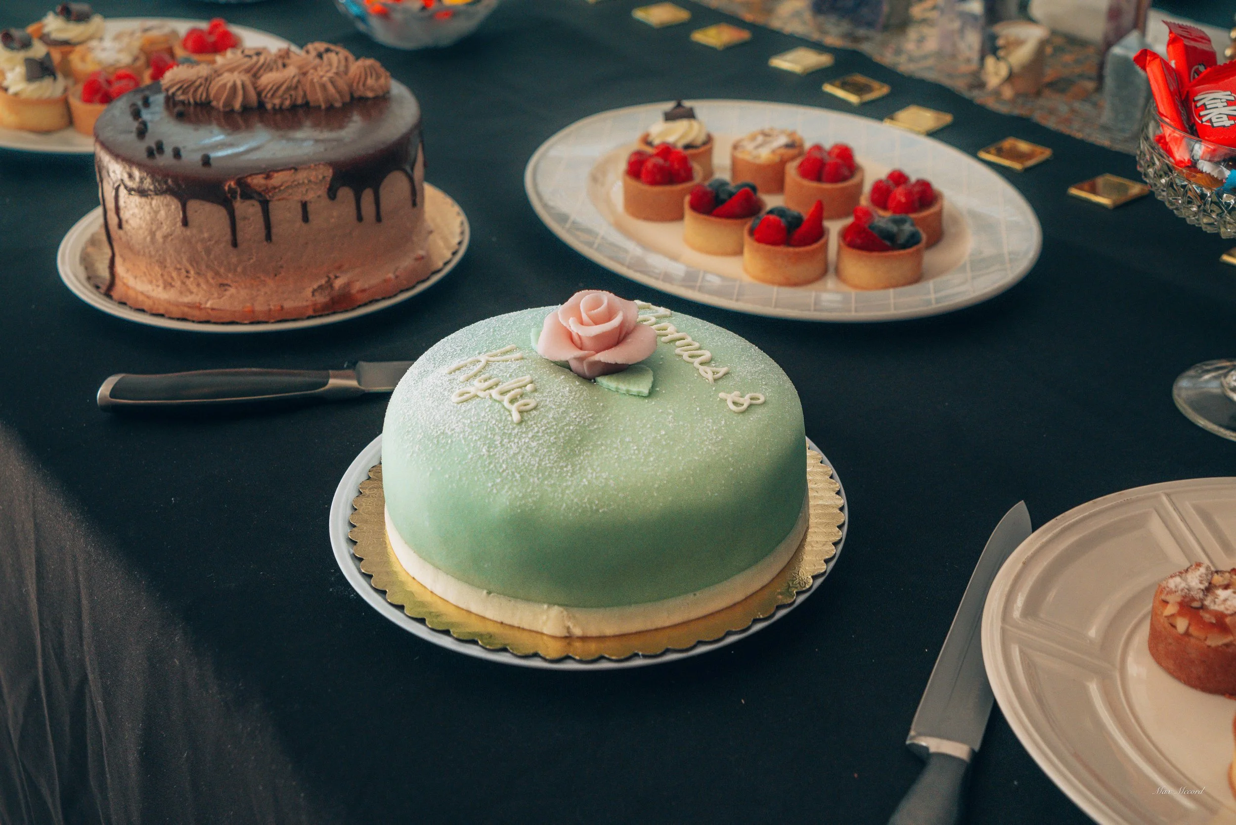 A green cake with a pink rose decoration on top, surrounded by other assorted cakes and desserts on a table.