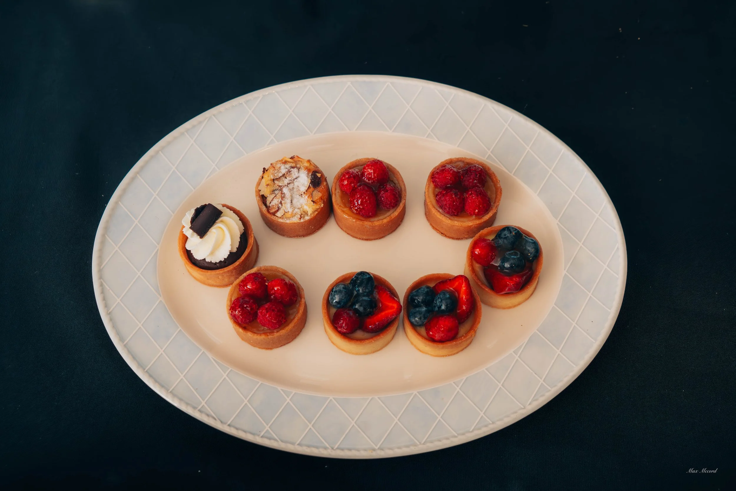 A white oval platter with ten mini fruit tarts, topped with assorted berries and whipped cream, placed on a dark table.