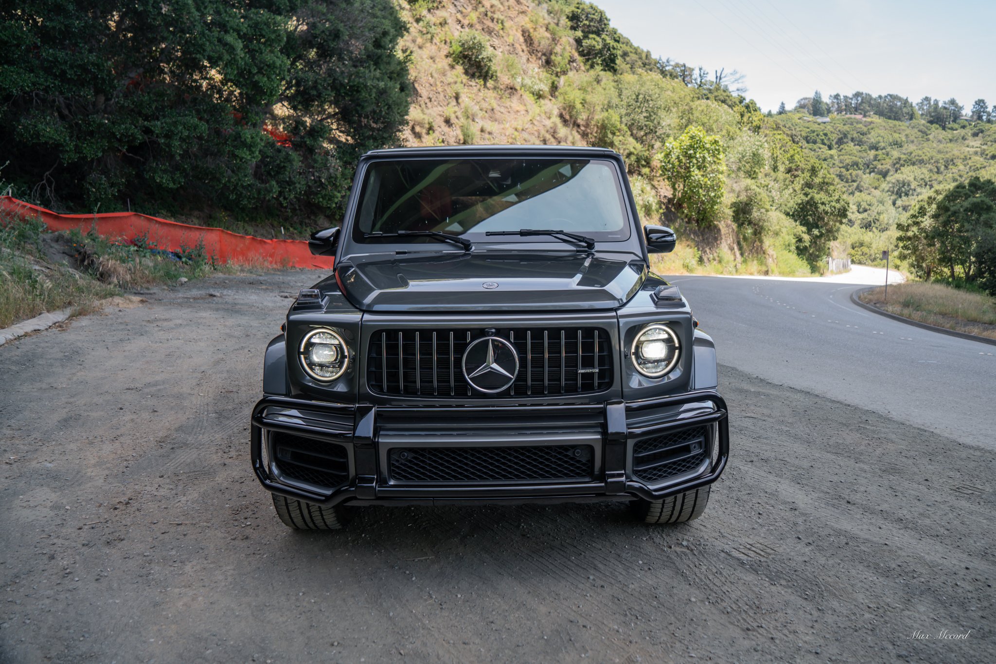 Front view of a black Mercedes-Benz G-Class SUV parked on a dirt road with a curving paved road and green hilly landscape in the background.