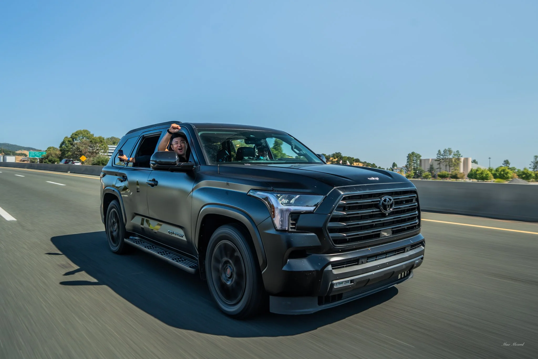 Black Toyota SUV driving on highway with a person inside celebrating and holding a phone, clear blue sky in the background.