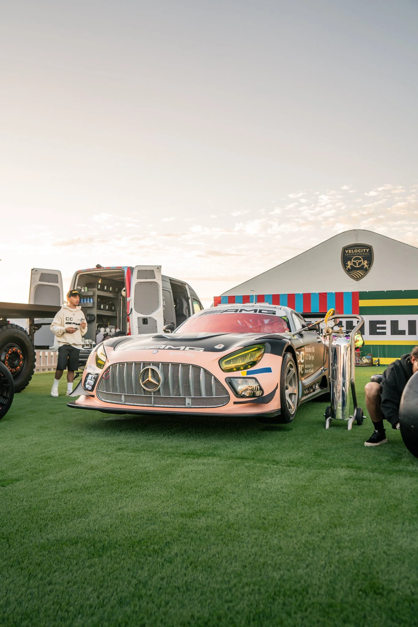 A Mercedes-Benz race car in a pit area with a large truck and a tent labeled 'Velocity' in the background, during sunset.