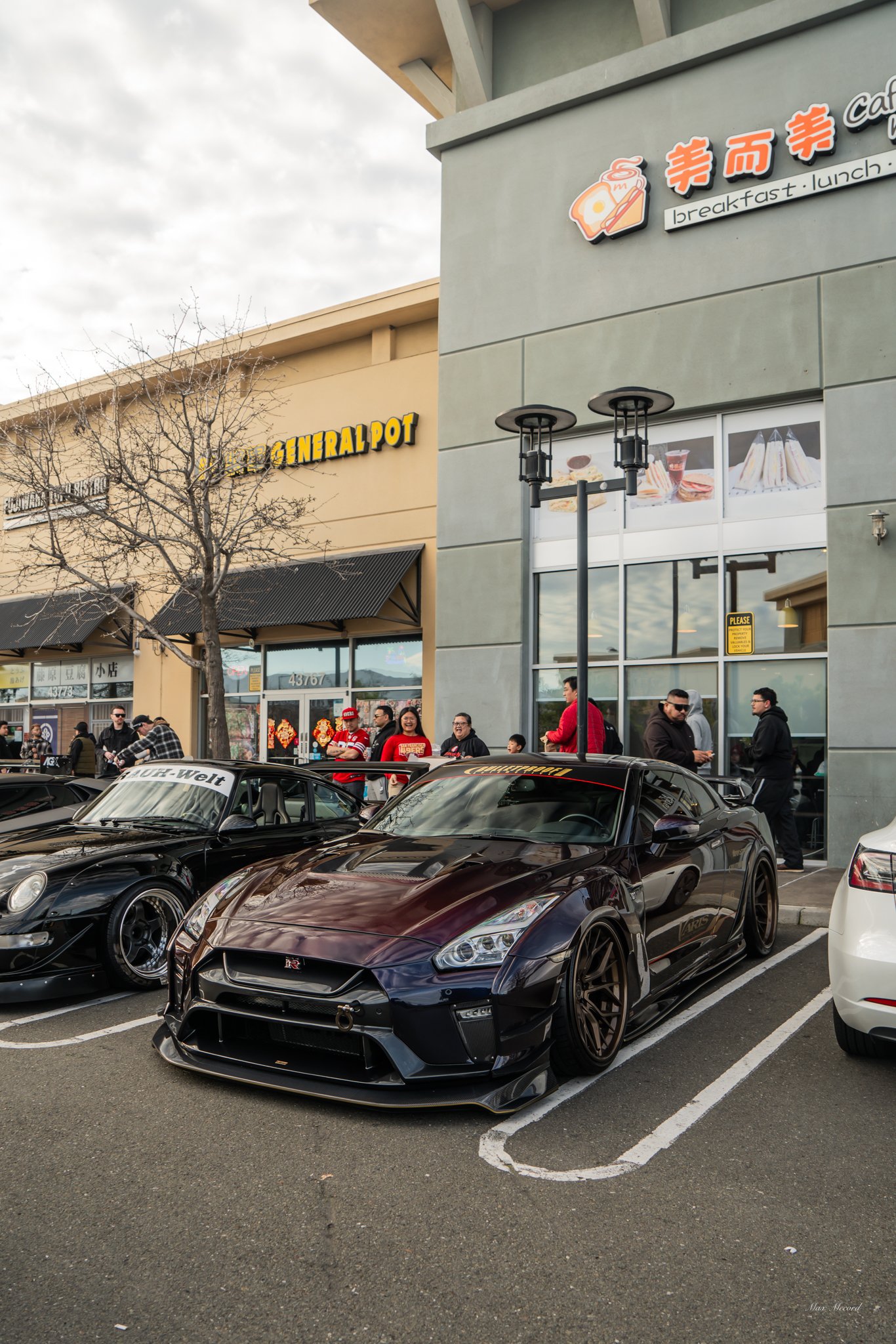 Black Nissan GT-R sports car parked in front of a coffee shop, with people standing outside and other cars parked nearby.
