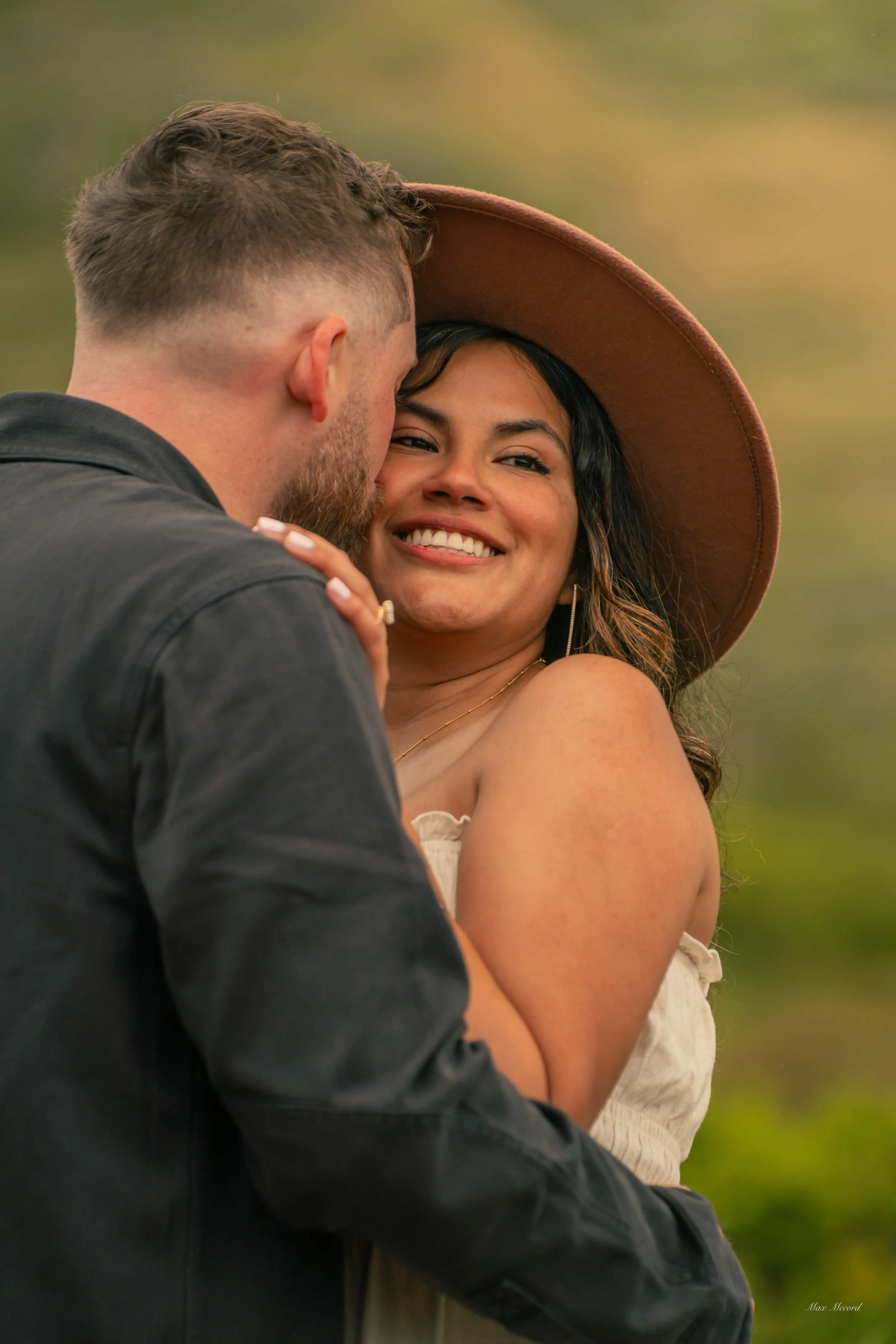 A woman with long dark hair and a large floppy hat smiling as a man leans in close, outside with blurred greenery in the background.