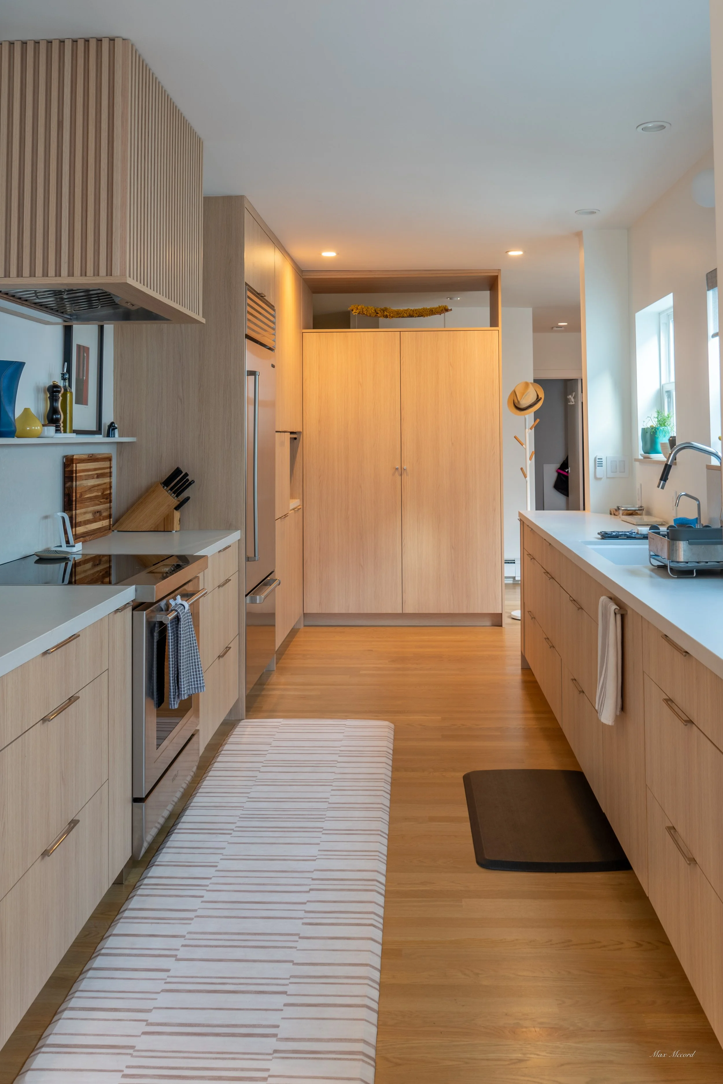 A modern kitchen with light wood cabinets, a white countertop, and a hardwood floor. The kitchen has a stove, refrigerator, and window with plants, with some decorative items and a hat hanging on a coat rack.